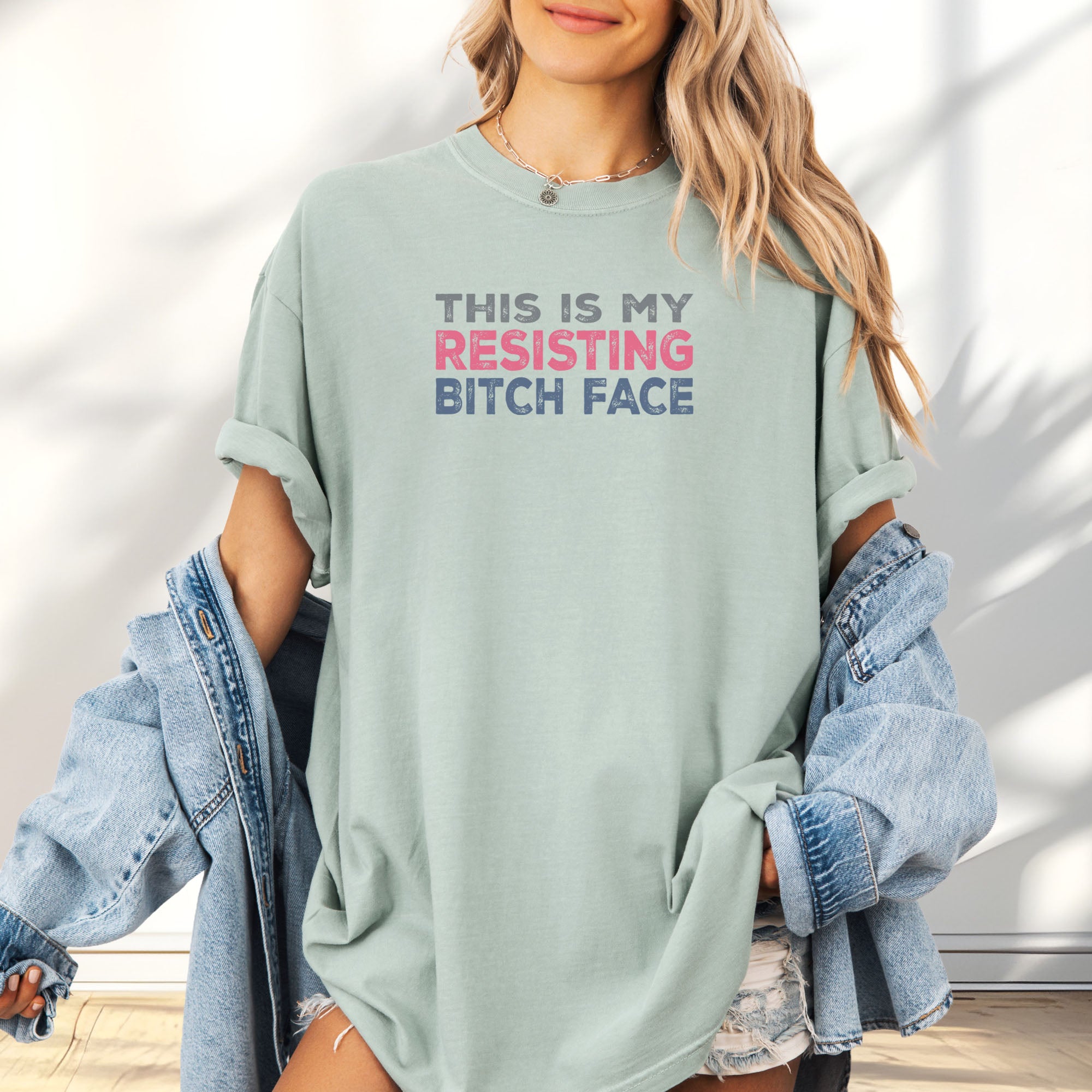 Female model wearing bay t-shirt with bold text that reads "This Is My Resisting Bitch Face" in gray, pink, and blue distressed font – a strong feminist and political protest shirt ideal for activism and making a bold statement.