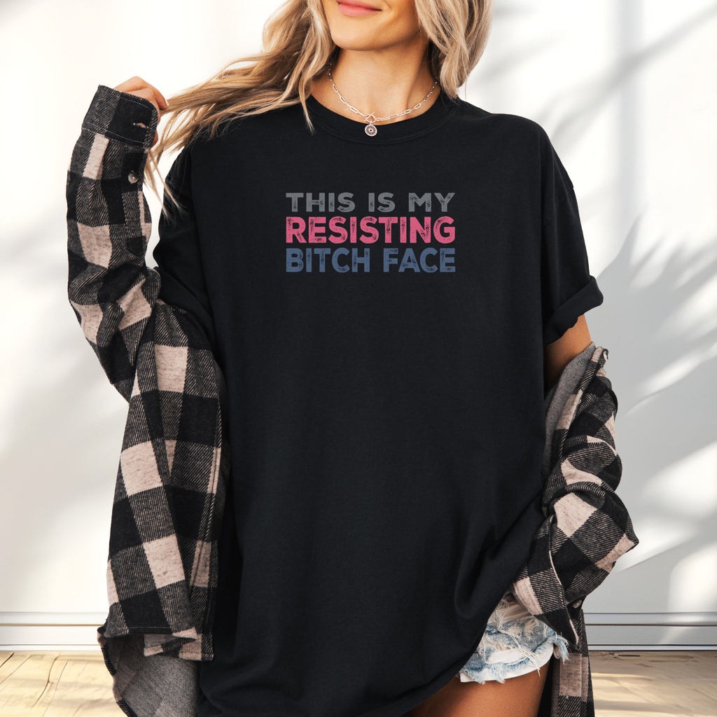 Female model wearing a black t-shirt with bold text that reads "This Is My Resisting Bitch Face" in gray, pink, and blue distressed font – a strong feminist and political protest shirt ideal for activism and making a bold statement.