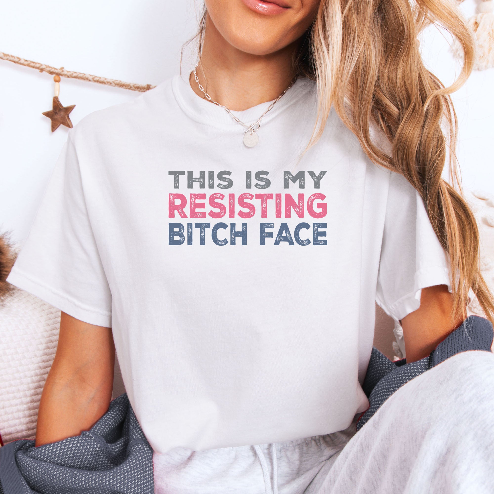 Female model wearing a white t-shirt with bold text that reads "This Is My Resisting Bitch Face" in gray, pink, and blue distressed font – a strong feminist and political protest shirt ideal for activism and making a bold statement.