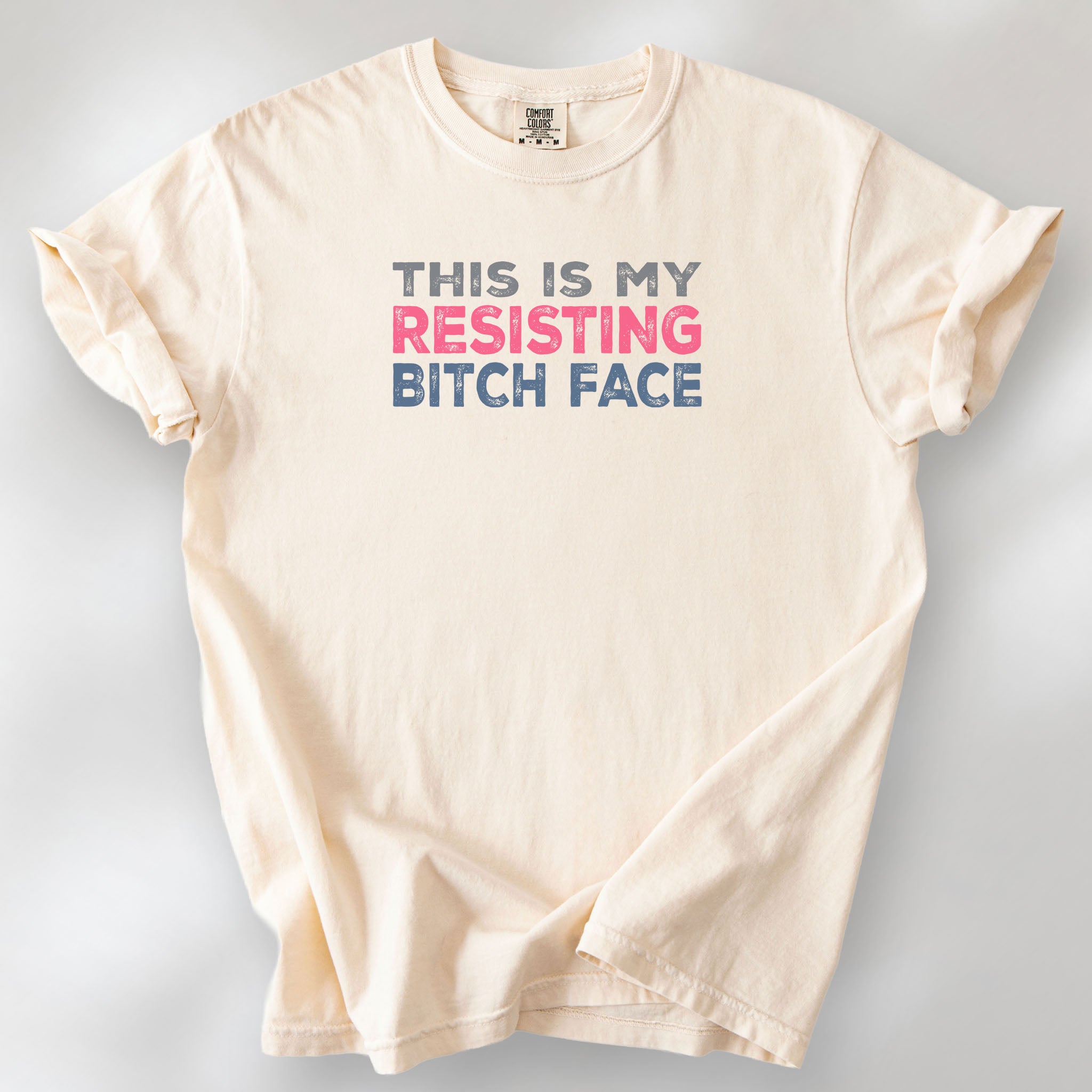 Ivory t-shirt with bold text that reads "This Is My Resisting Bitch Face" in gray, pink, and blue distressed font – a strong feminist and political protest shirt ideal for activism and making a bold statement.