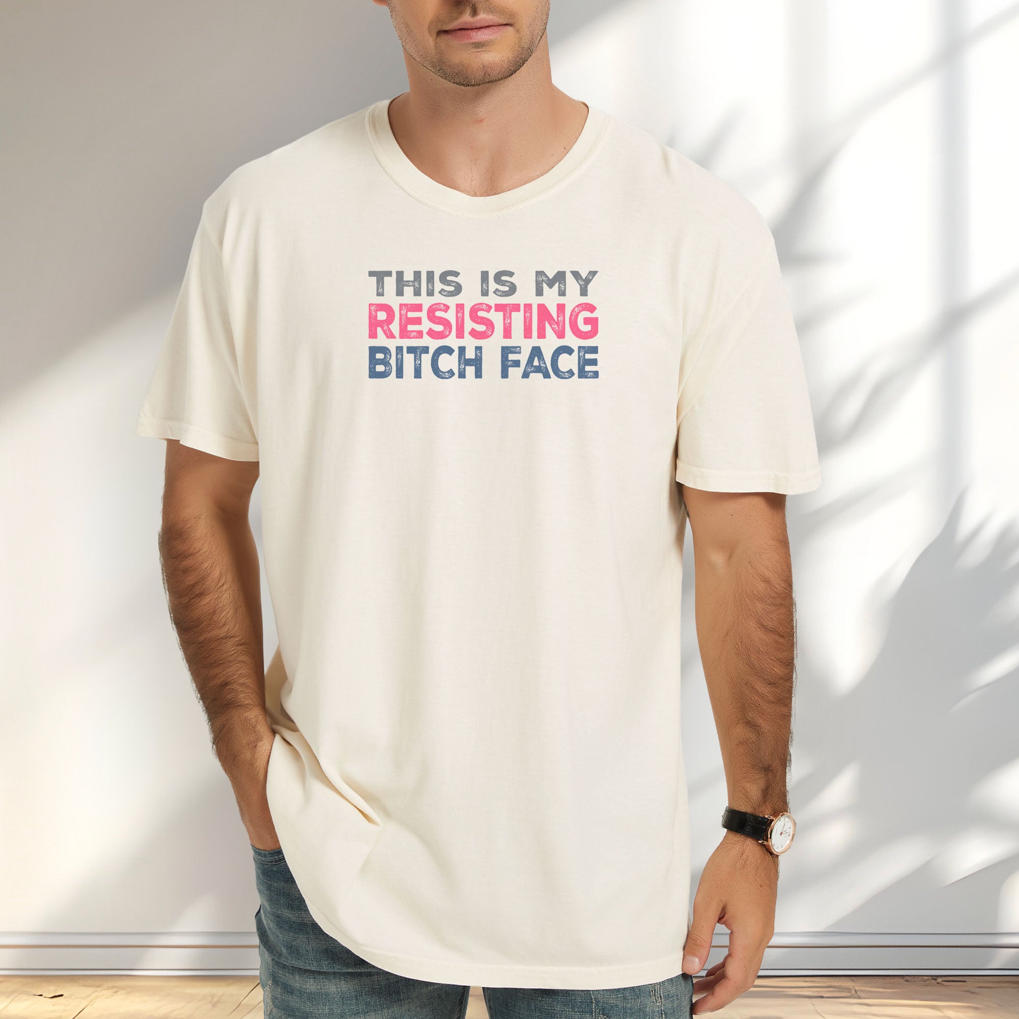 Male model wearing ivory t-shirt with bold text that reads "This Is My Resisting Bitch Face" in gray, pink, and blue distressed font – a strong feminist and political protest shirt ideal for activism and making a bold statement.