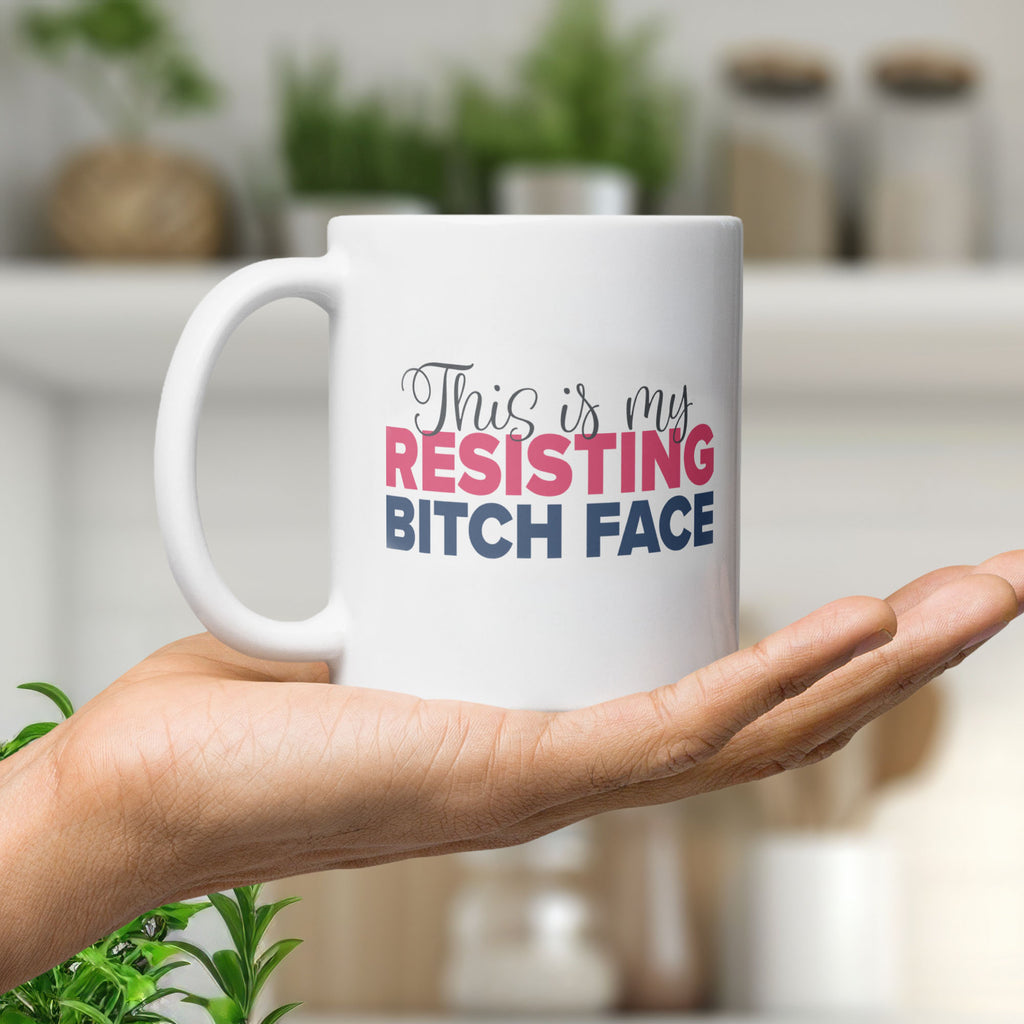 Glossy white ceramic coffee mug resting on a person's palm in a bright kitchen, with a graphic that reads "This is my resisting bitch face" in stylized text.