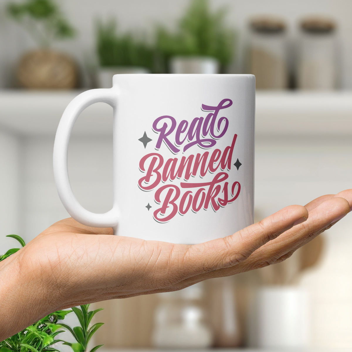 White ceramic coffee mug with stylized text graphic that reads "read banned books" in purple and red text, held in the palm of a hand in a kitchen.