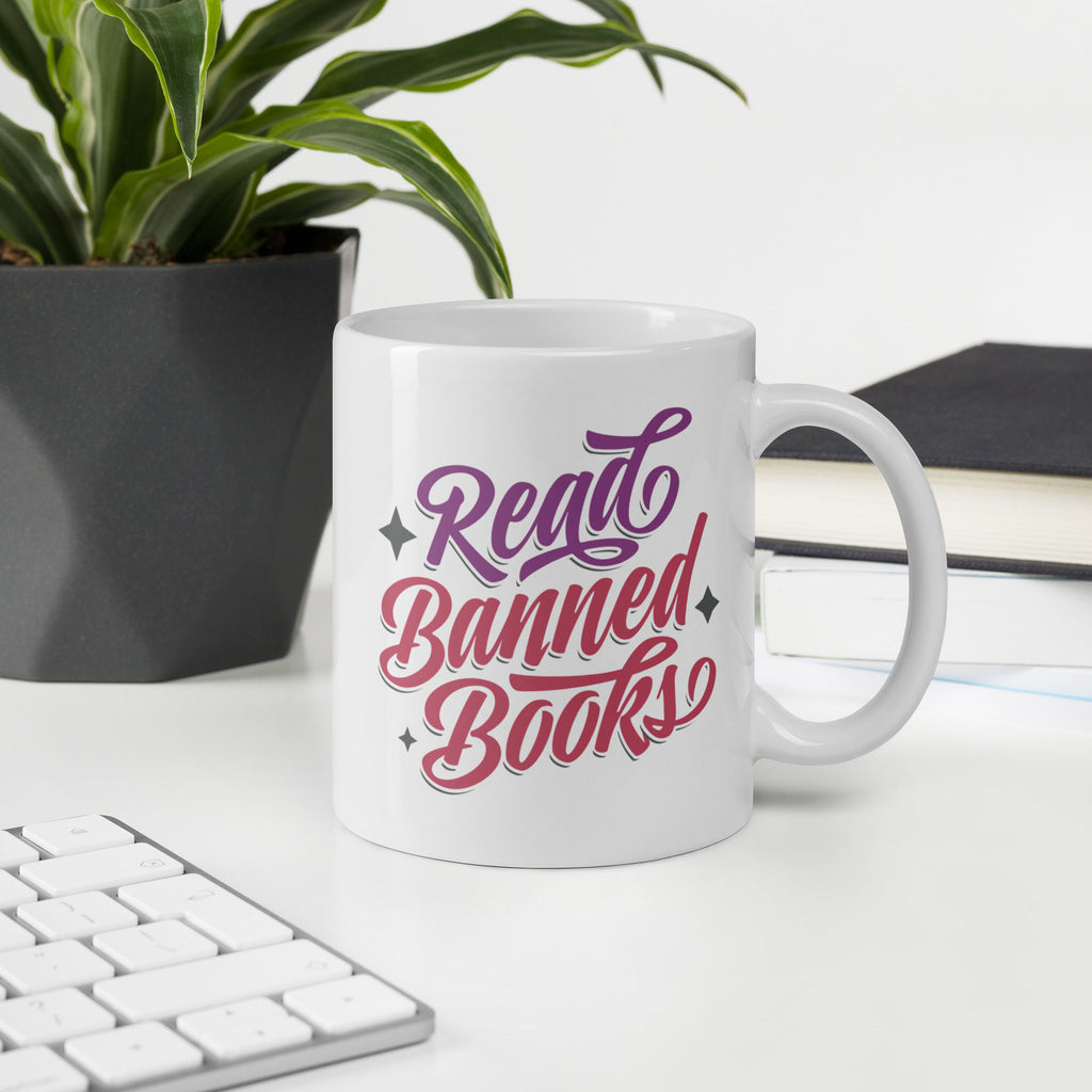 White ceramic coffee mug with stylized text graphic that reads "read banned books" in purple and red text, on a desk with a book, keyboard, and plant.