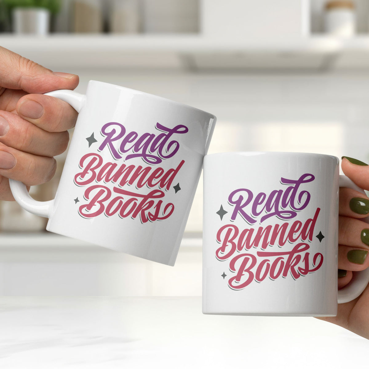 Two white ceramic coffee mugs with stylized text graphic that reads "read banned books" in purple and red text, held by two hands, being clinked together in a white kitchen.