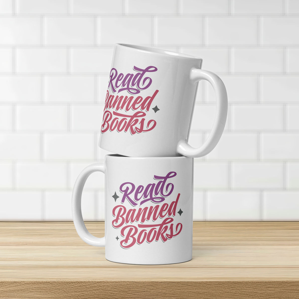 Two stacked white ceramic coffee mugs with stylized text graphic that reads "read banned books" in purple and red text, on a kitchen counter.