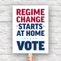 Protest sign reading “Regime Change Starts at Home — Vote” in red and blue political typography, held on a wooden stick against a brick wall background.