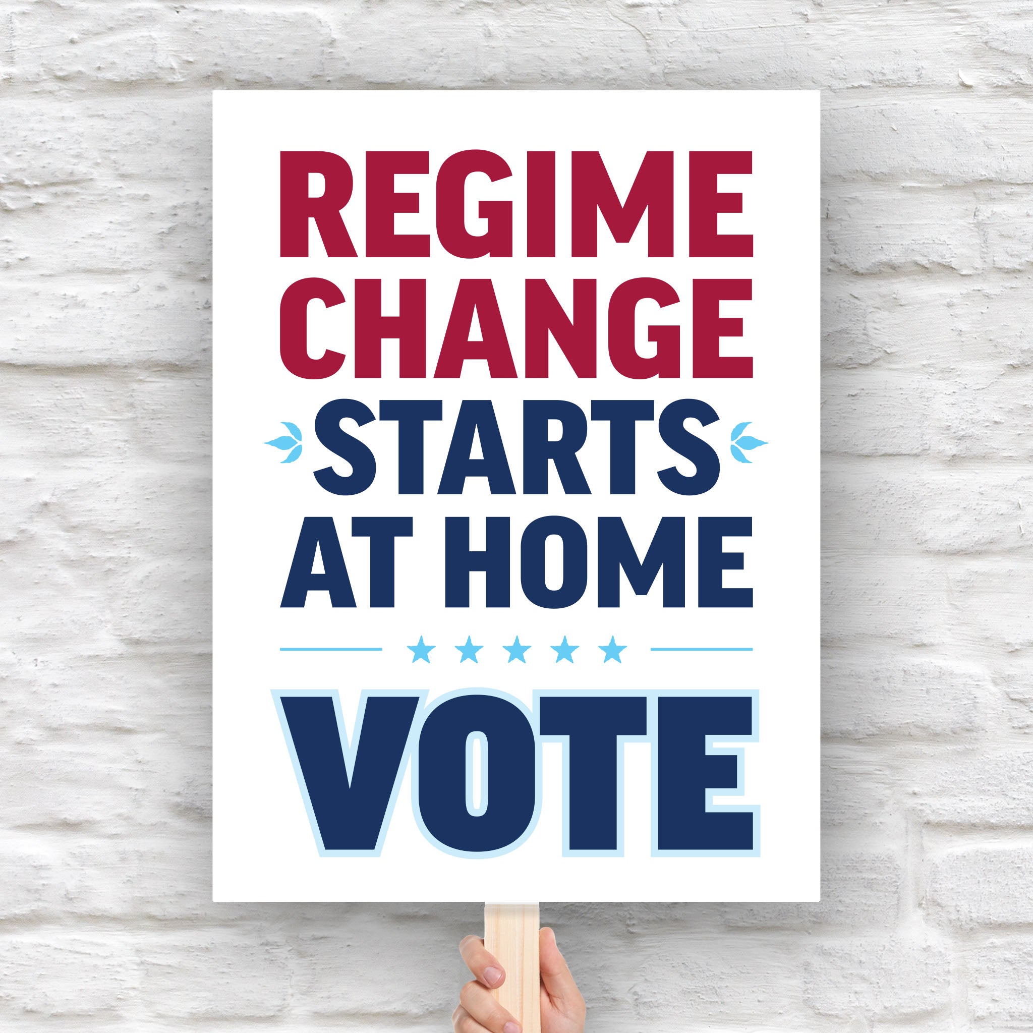 Protest sign reading “Regime Change Starts at Home — Vote” in red and blue political typography, held on a wooden stick against a brick wall background.