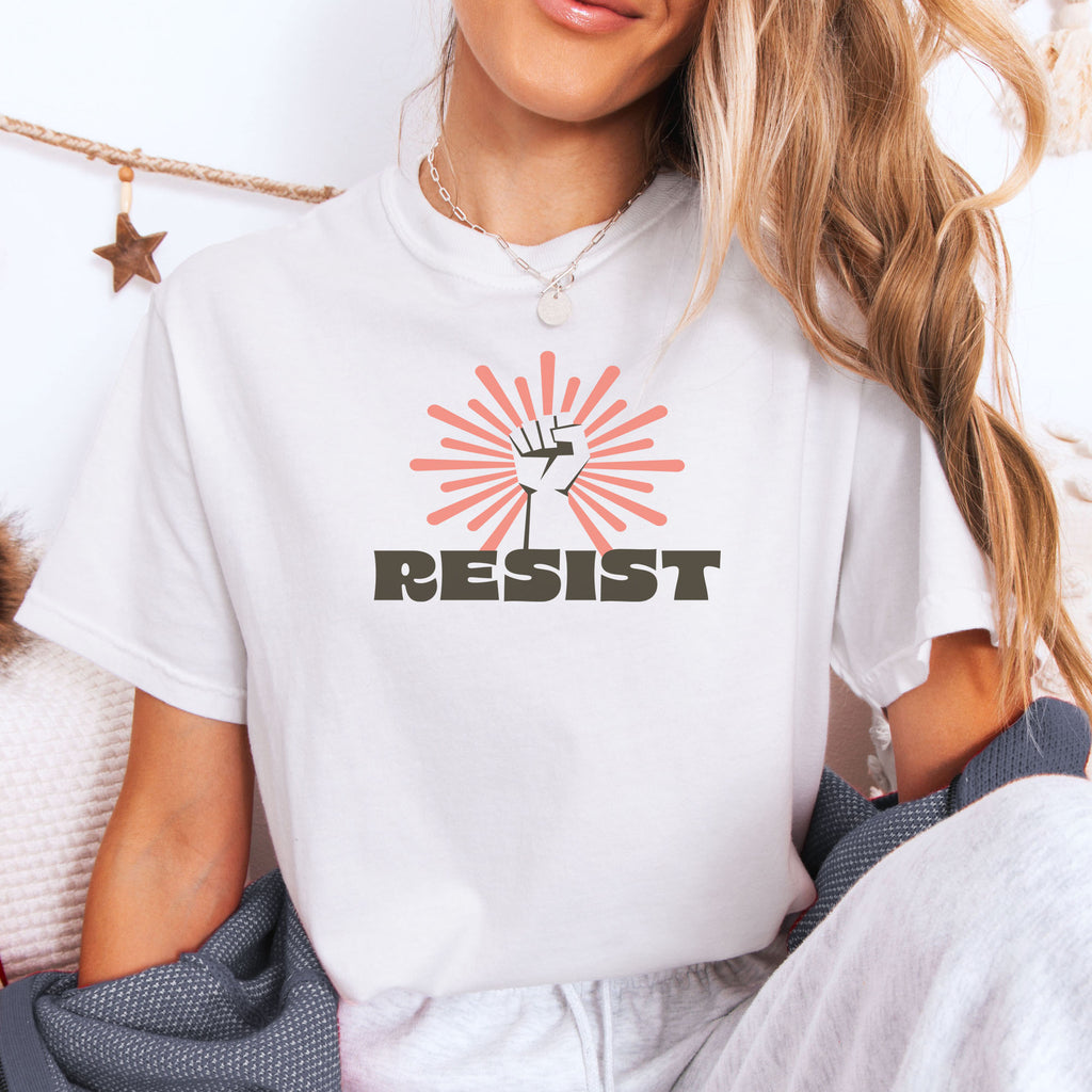 Female model wearing white t-shirt with retro style "resist" text, raised fist, and pinkish burst behind the fist.