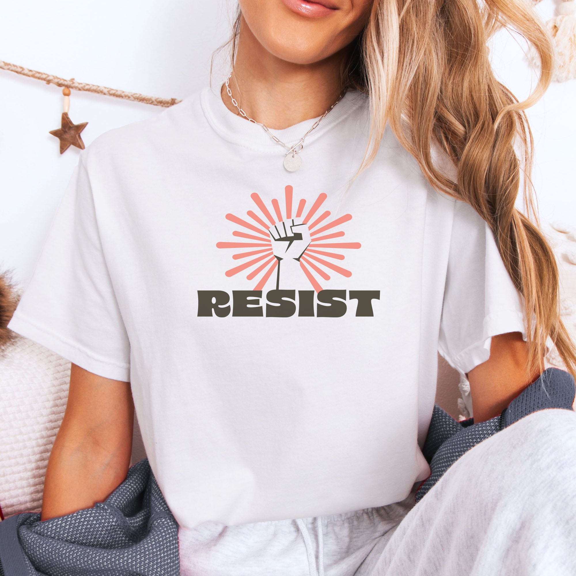 Female model wearing white t-shirt with retro style "resist" text, raised fist, and pinkish burst behind the fist.