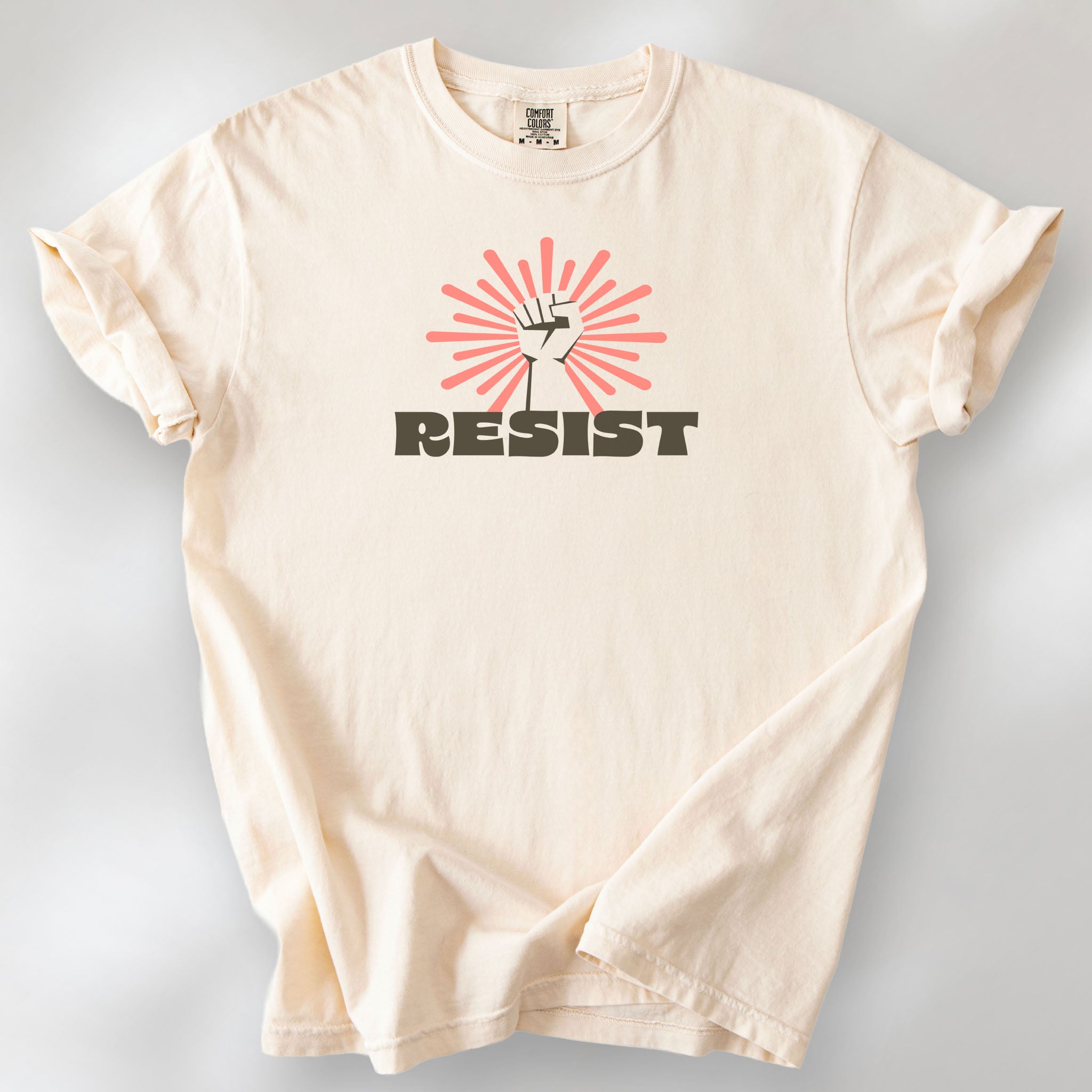 Ivory t-shirt with retro style "resist" text, raised fist, and pinkish burst behind the fist.