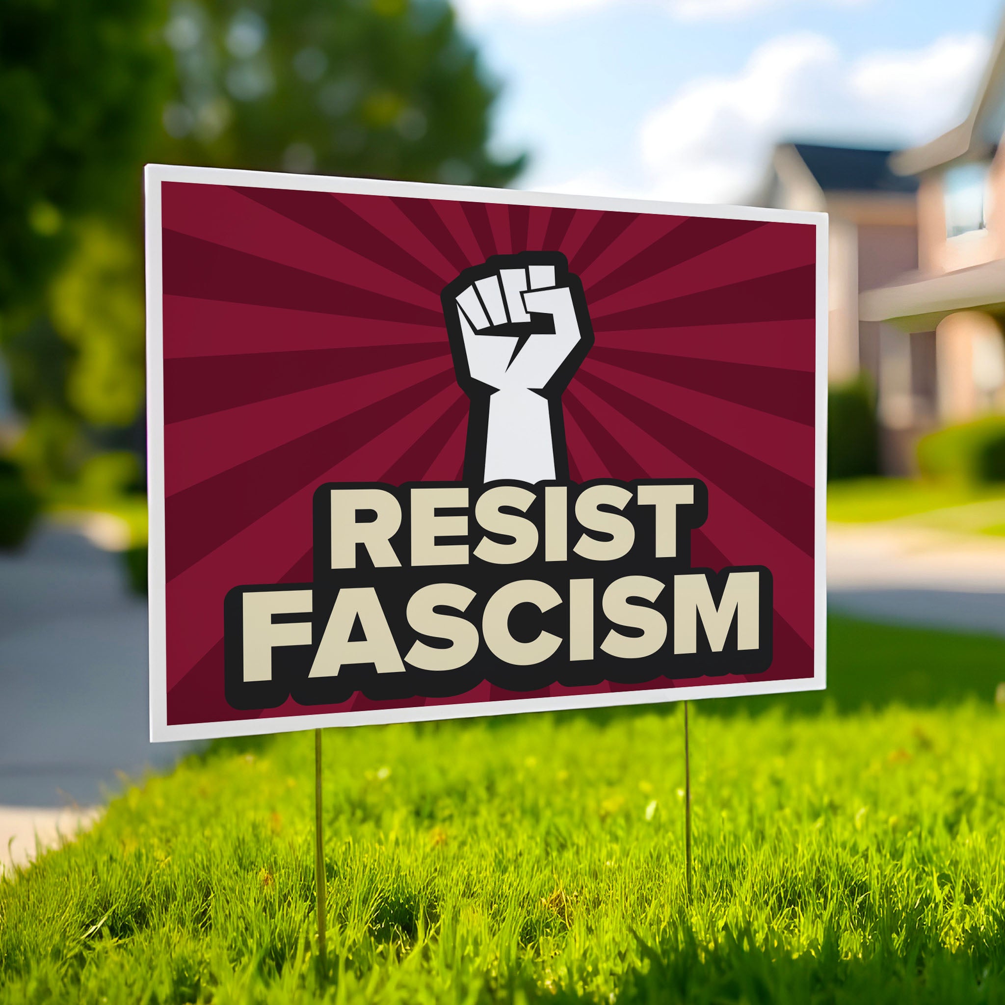 Yard sign with red burst background and white raised fist above text that reads "Resist fascism" in all uppercase letters, planted in a green lawn.