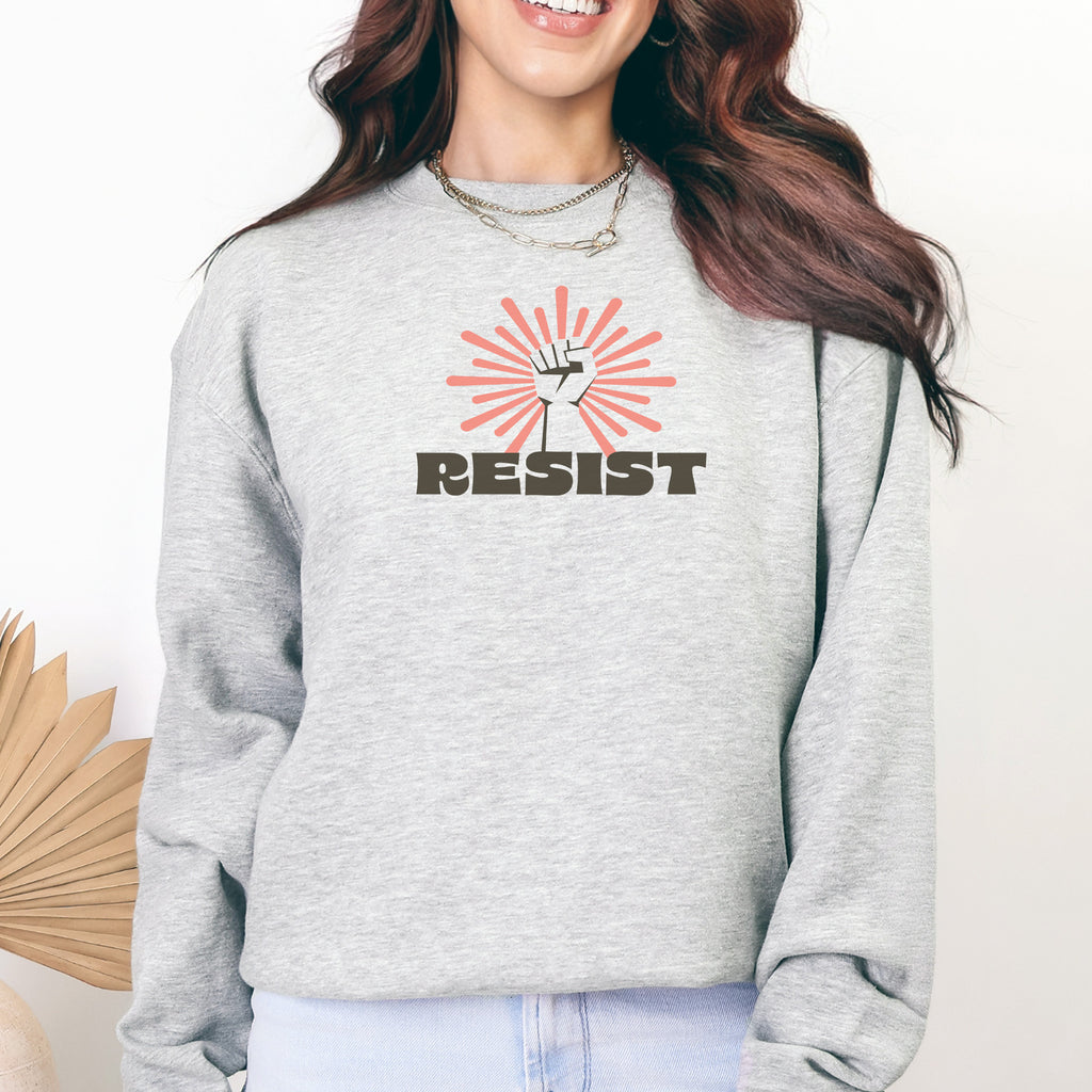 Female model wearing grey sweatshirt with retro style "resist" text, raised fist, and pinkish burst behind the fist.