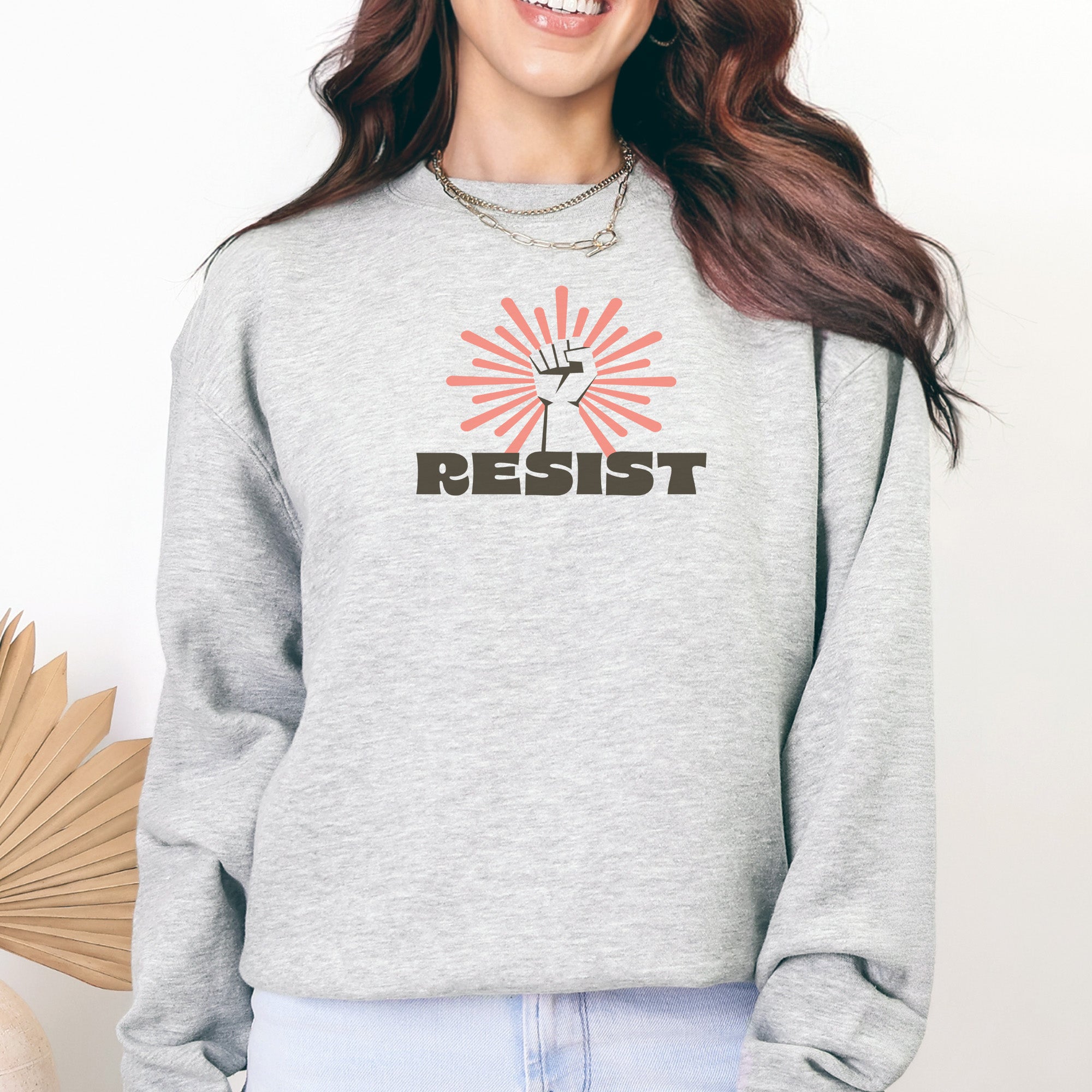 Female model wearing grey sweatshirt with retro style "resist" text, raised fist, and pinkish burst behind the fist.
