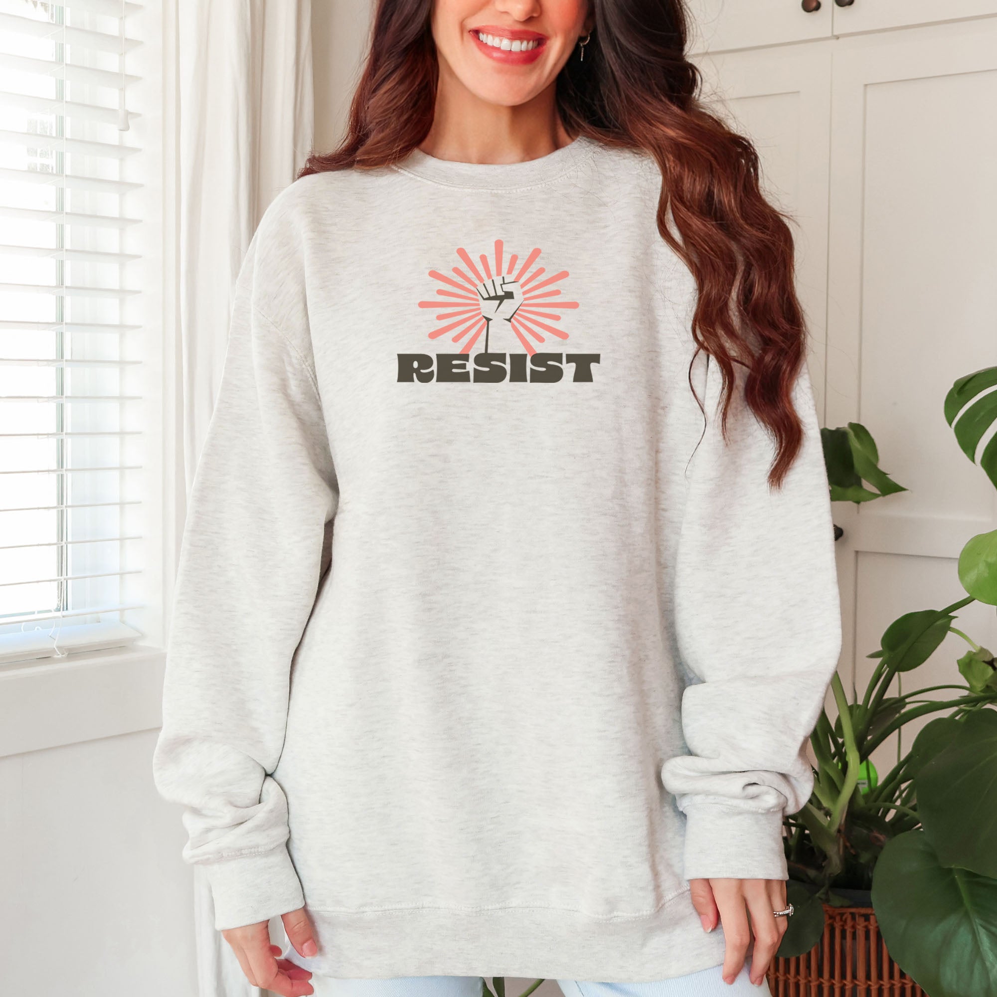Female model wearing oatmeal sweatshirt with retro style "resist" text, raised fist, and pinkish burst behind the fist.