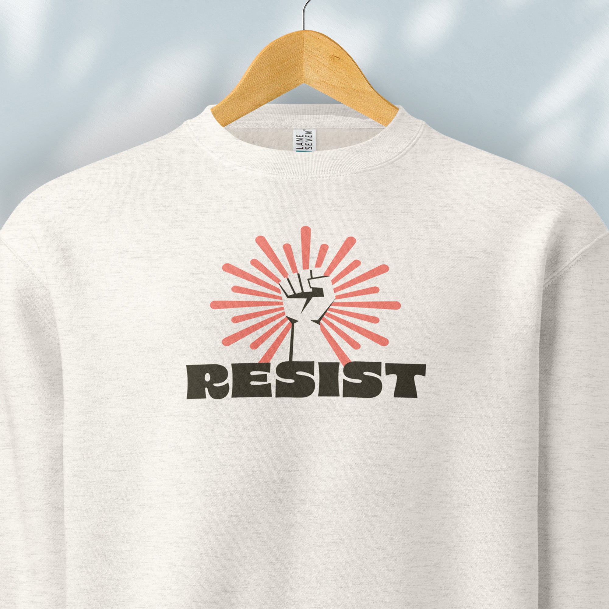 Hanging oatmeal sweatshirt with retro style "resist" text, raised fist, and pinkish burst behind the fist.