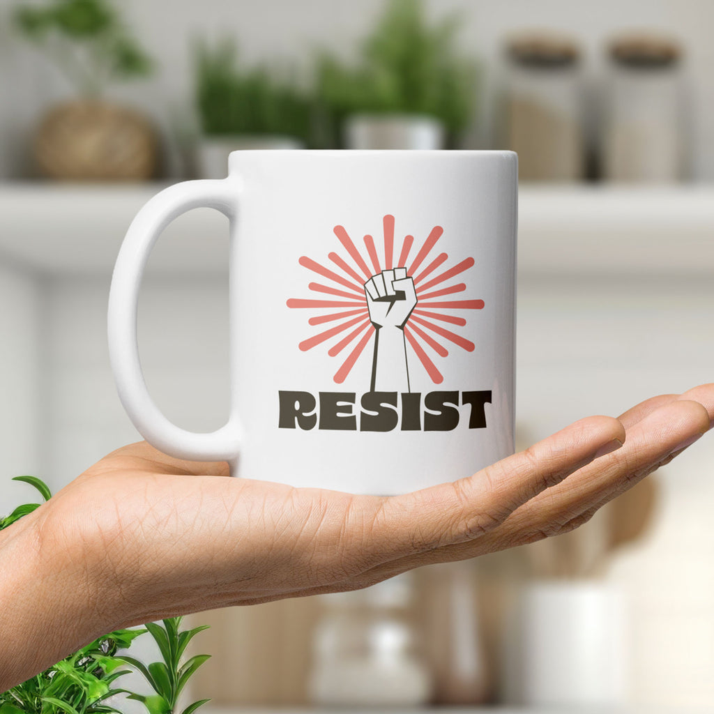 Coffee mug held in the palm of a hand. Mug has a raised fist graphic in front of a coral burst, and bold retro-styled text that says "resist."