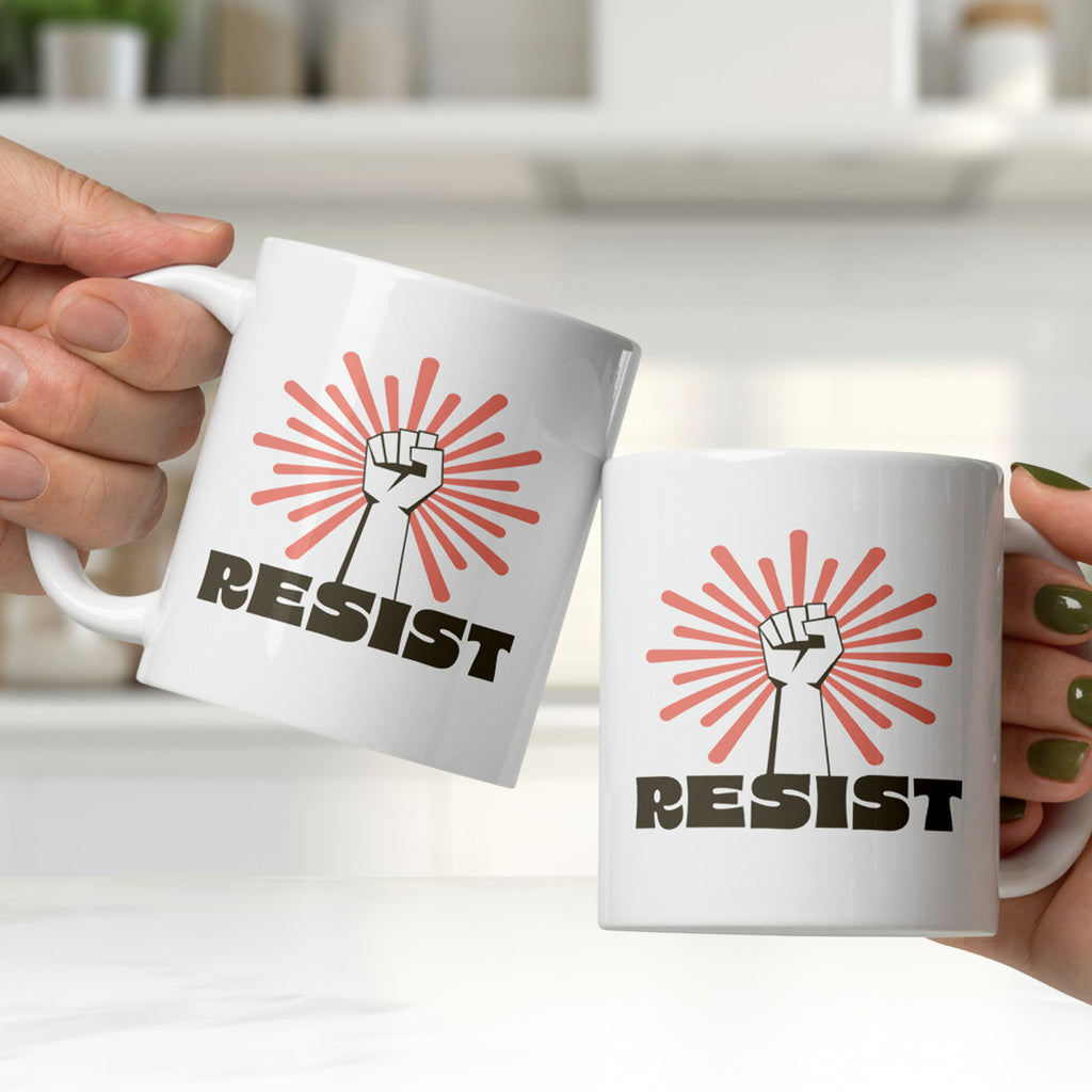 Two ceramin mugs held by people clinking them together in a bright white kitchen. Mug has a raised fist graphic in front of a coral burst, and bold retro-styled text that says "resist."