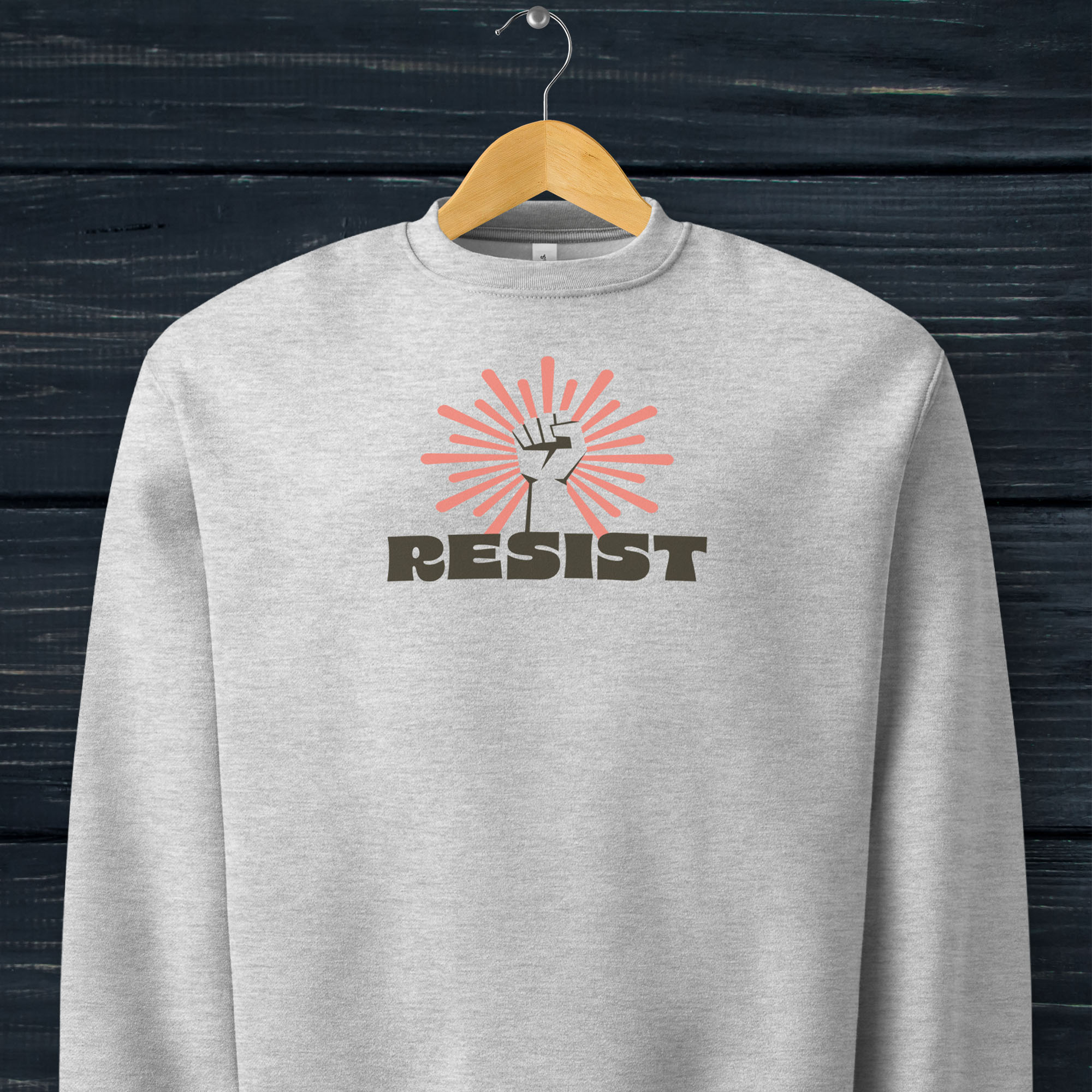 Athletic heather grey sweatshirt with "resist" text in bold retro style, below a raised fist with a coral color burst behind it, hanging on a hanger.