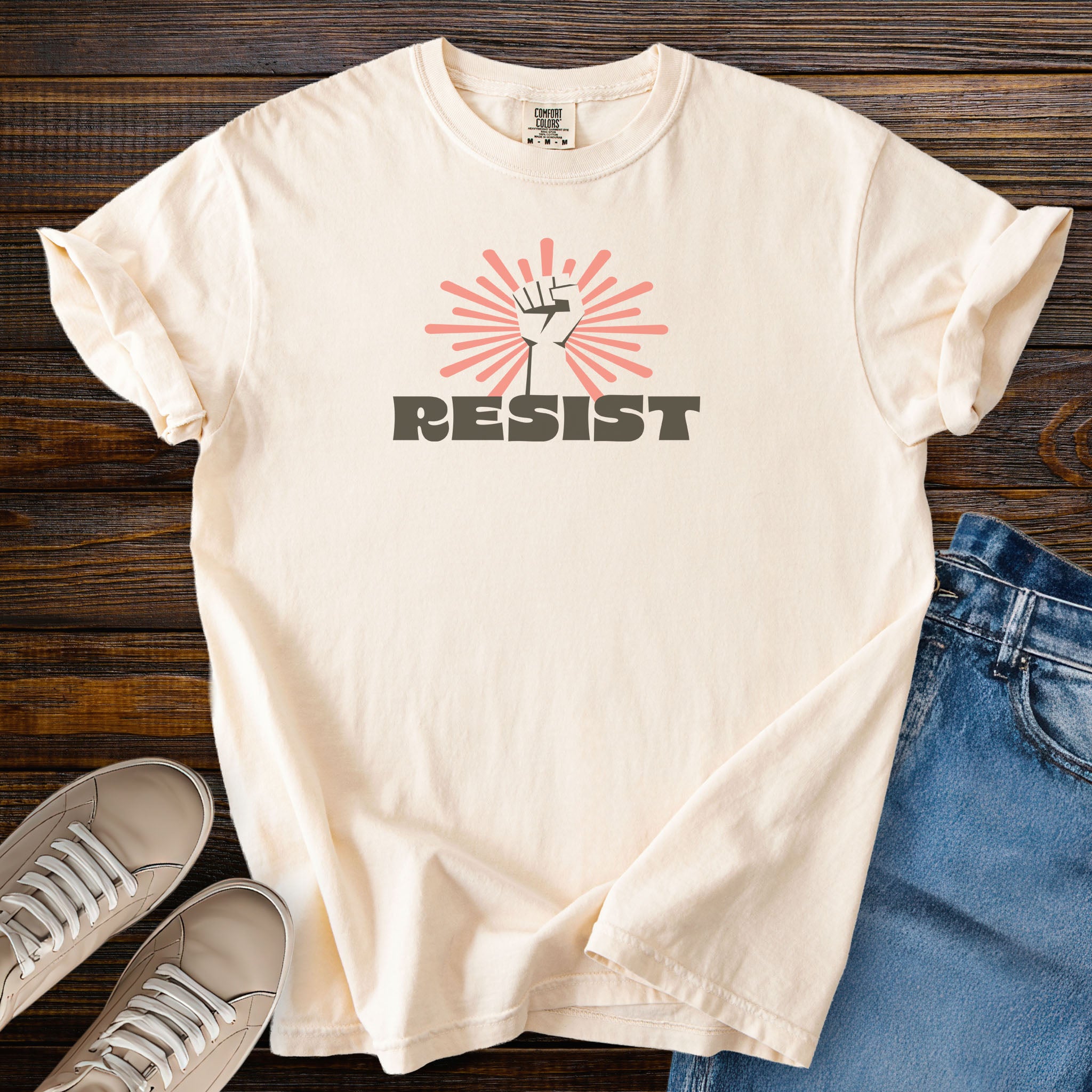 Ivory color t-shirt with retro-styled 'resist' text below a raised fist on a wood surface with jeans and sneakers.