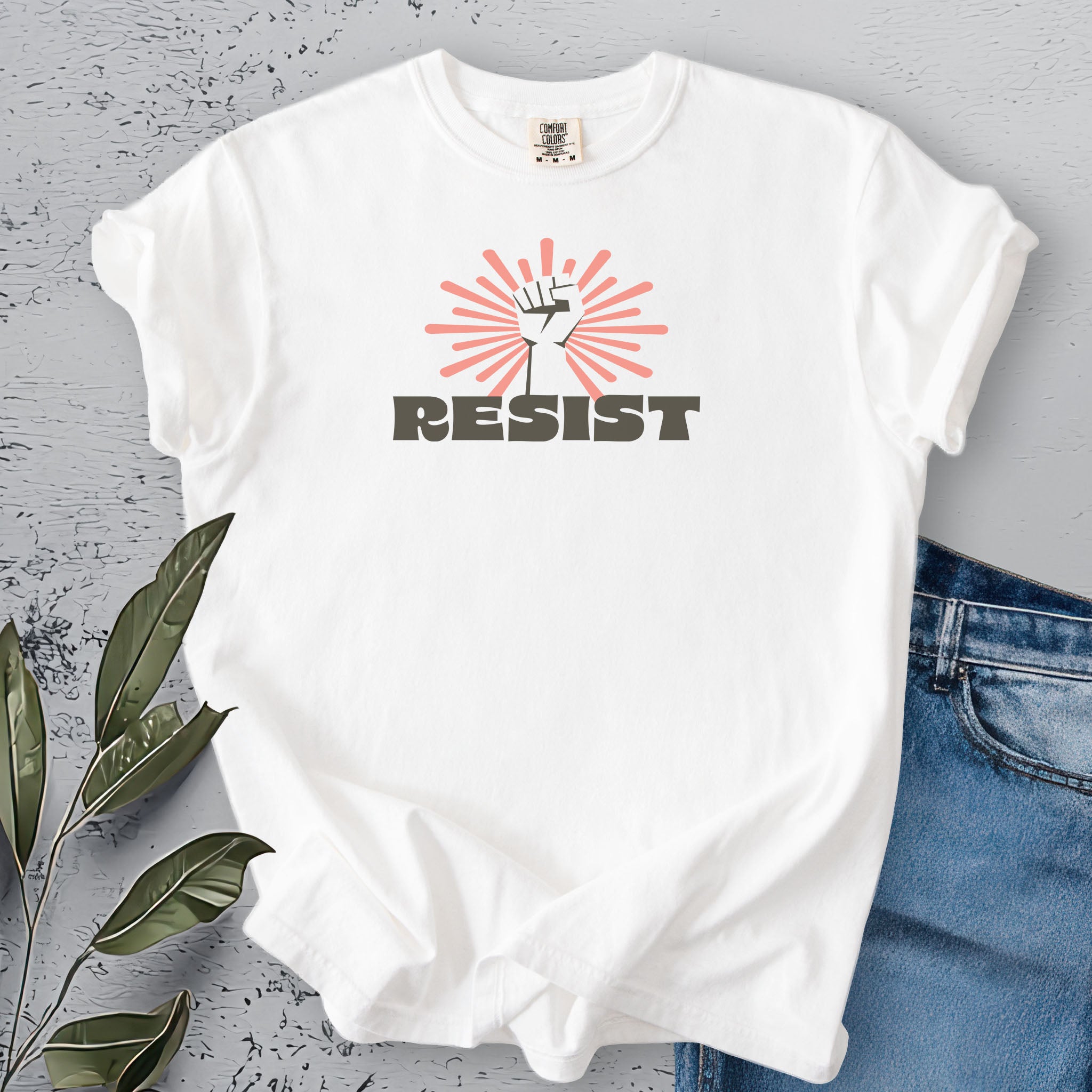 White t-shirt with retro-styled 'resist' text below a raised fist on a gray surface with leaves and blue jeans.