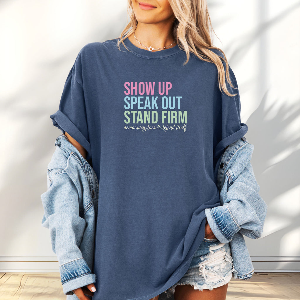 Female model wearing oversized denim t-shirt with bright pink, blue and green bold text that says "show up, speak out, stand firm" and subtle grey script text below that reads "democracy doesn't defend itself."