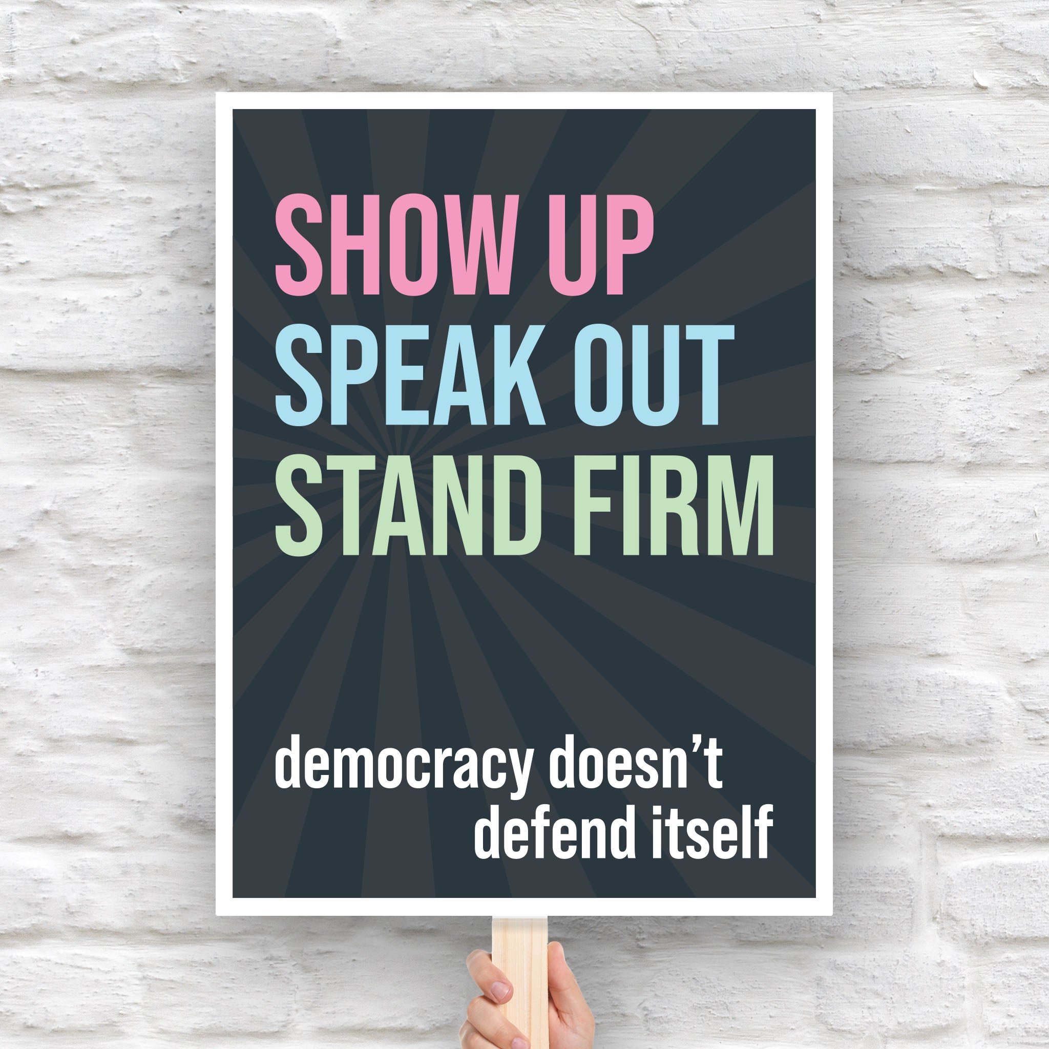Downloadable, printable protest sign with dark background with subtle burst, bold colorful text that reads "show up, speak out, stand firm", then white bold text that reads "democracy doesn't defend itself."