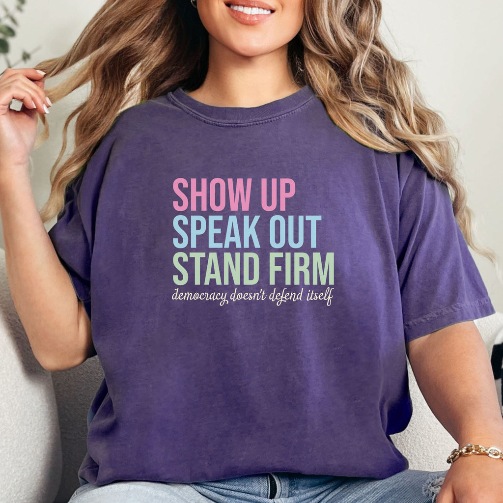 Female model wearing grape t-shirt with bright pink, blue and green bold text that says "show up, speak out, stand firm" and subtle grey script text below that reads "democracy doesn't defend itself."