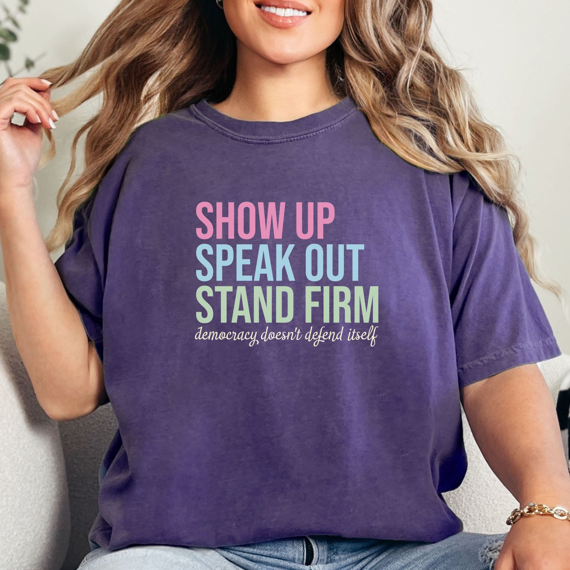 Female model wearing grape t-shirt with bright pink, blue and green bold text that says "show up, speak out, stand firm" and subtle grey script text below that reads "democracy doesn't defend itself."