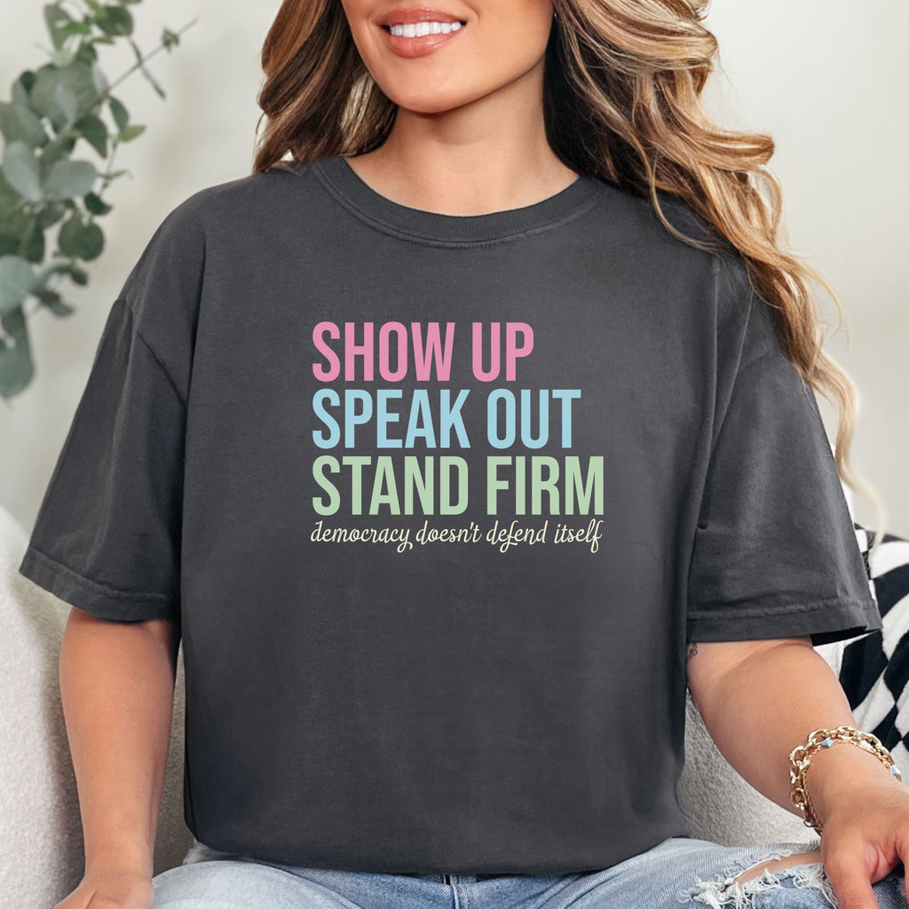 Female model wearing graphite t-shirt with bright pink, blue and green bold text that says "show up, speak out, stand firm" and subtle grey script text below that reads "democracy doesn't defend itself."