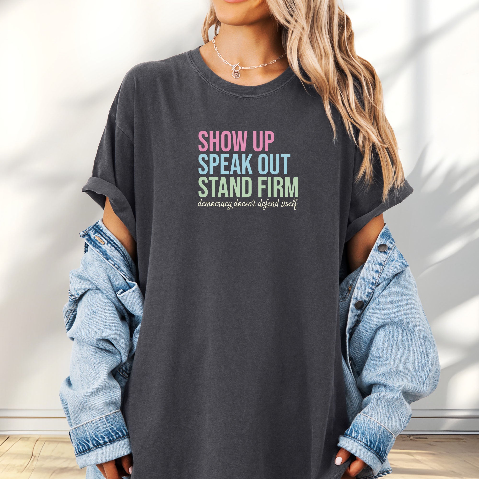 Female model wearing oversized pepper t-shirt with bright pink, blue and green bold text that says "show up, speak out, stand firm" and subtle grey script text below that reads "democracy doesn't defend itself."