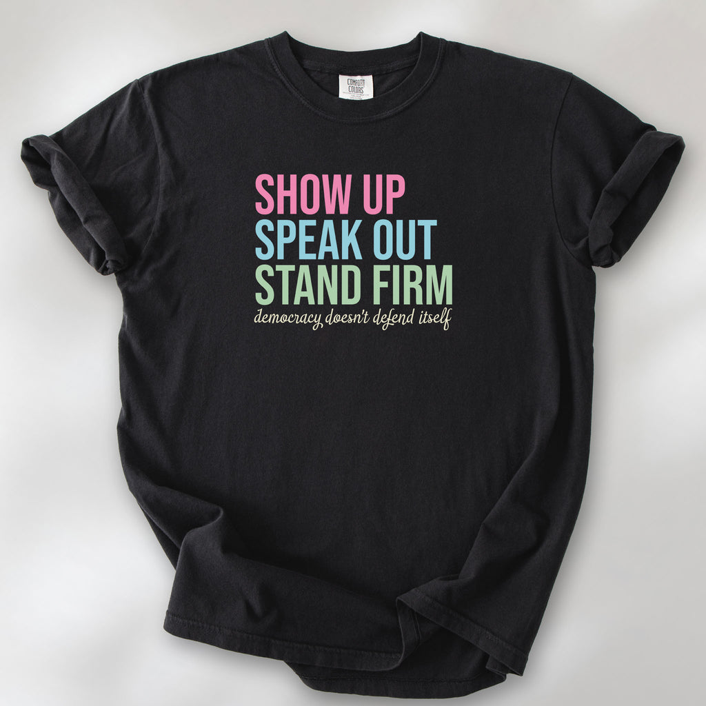 Black t-shirt with bright pink, blue and green bold text that says "show up, speak out, stand firm" and subtle grey script text below that reads "democracy doesn't defend itself."
