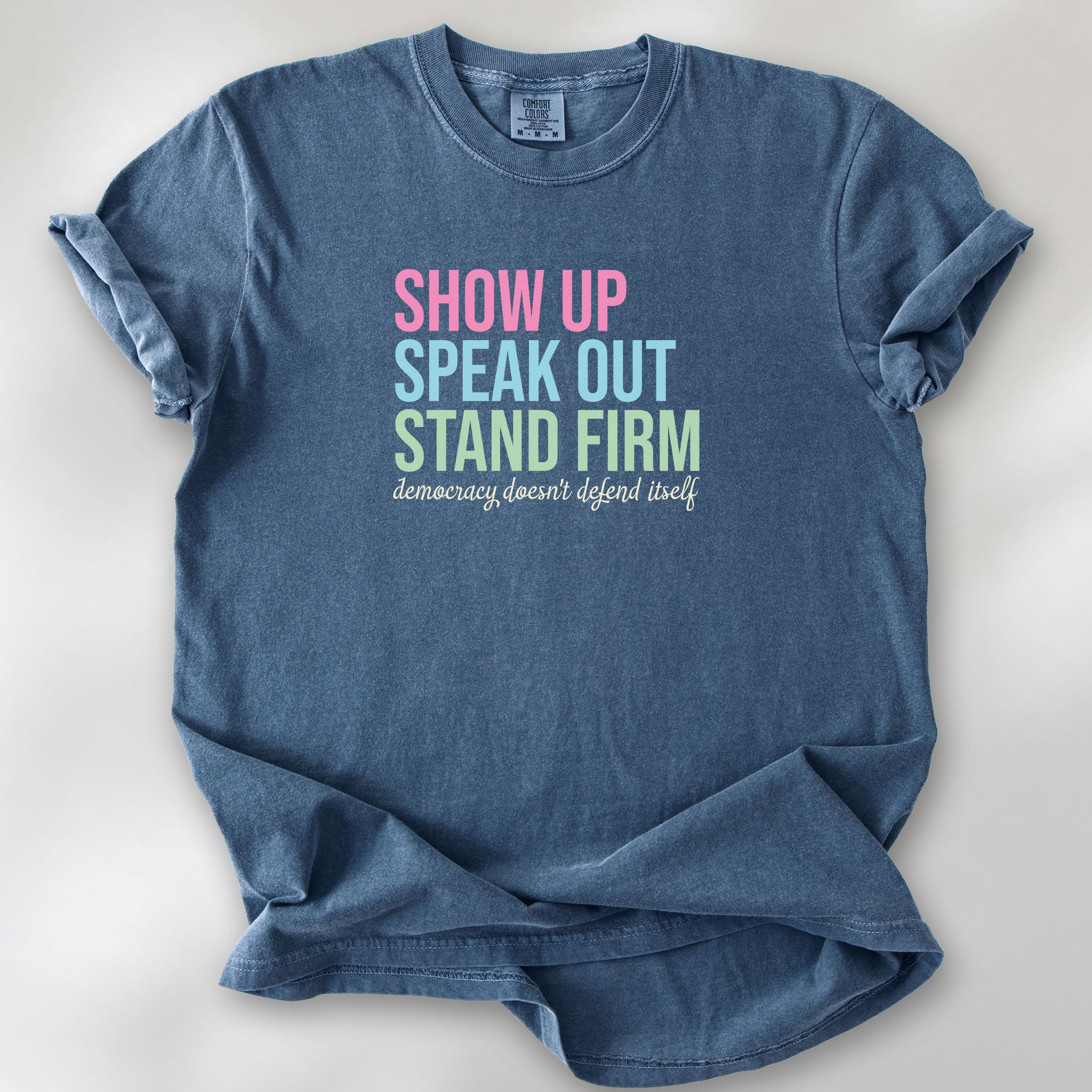 Denim t-shirt with bright pink, blue and green bold text that says "show up, speak out, stand firm" and subtle grey script text below that reads "democracy doesn't defend itself."