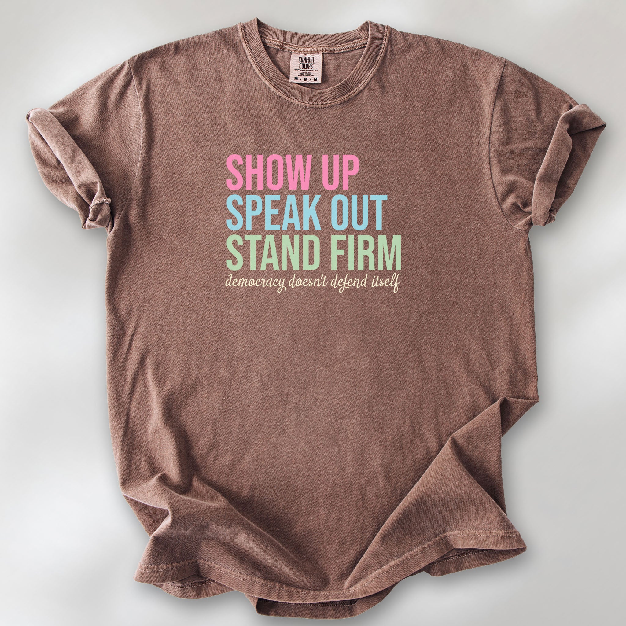 Espresso t-shirt with bright pink, blue and green bold text that says "show up, speak out, stand firm" and subtle grey script text below that reads "democracy doesn't defend itself."