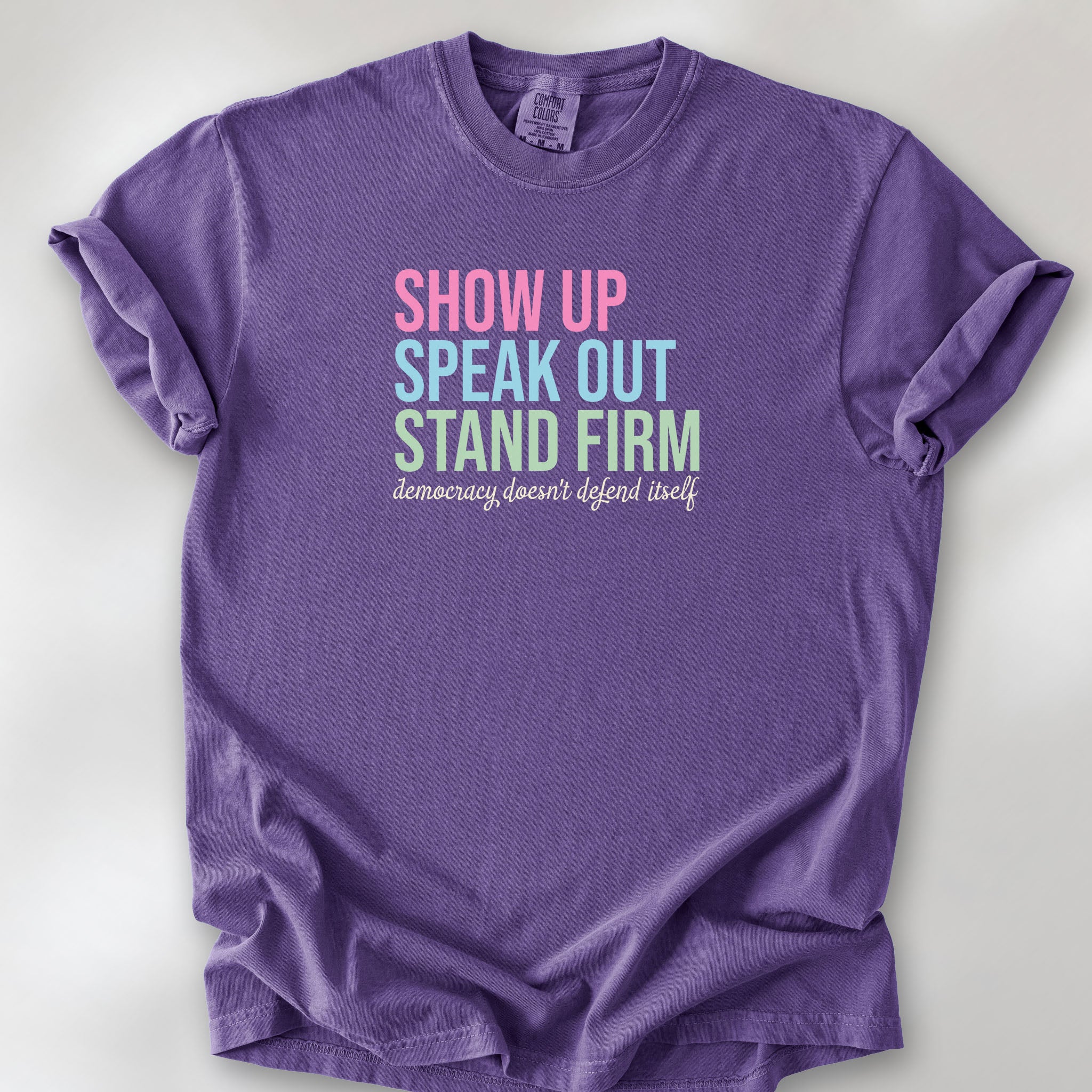 Grape t-shirt with bright pink, blue and green bold text that says "show up, speak out, stand firm" and subtle grey script text below that reads "democracy doesn't defend itself."