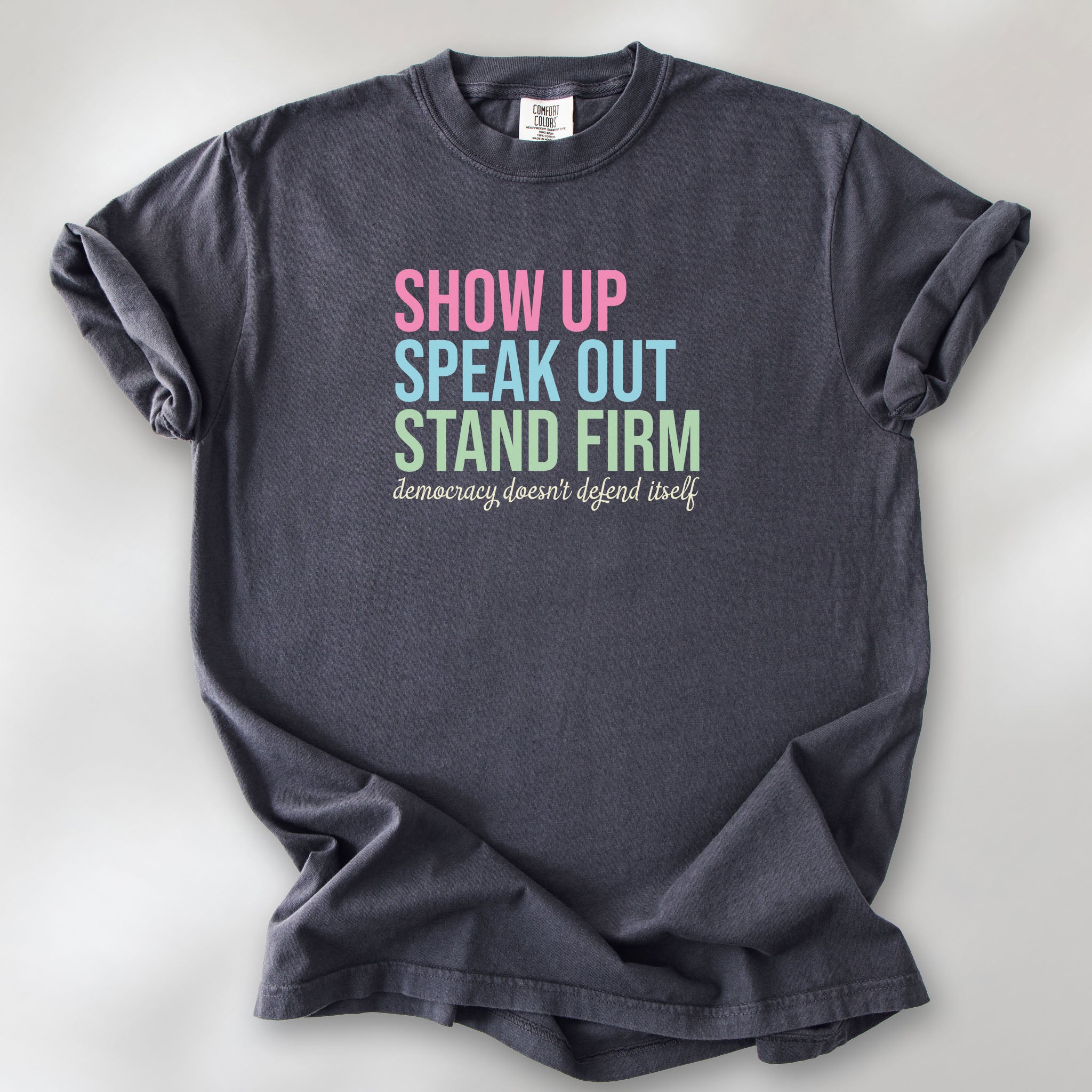Graphite t-shirt with bright pink, blue and green bold text that says "show up, speak out, stand firm" and subtle grey script text below that reads "democracy doesn't defend itself."