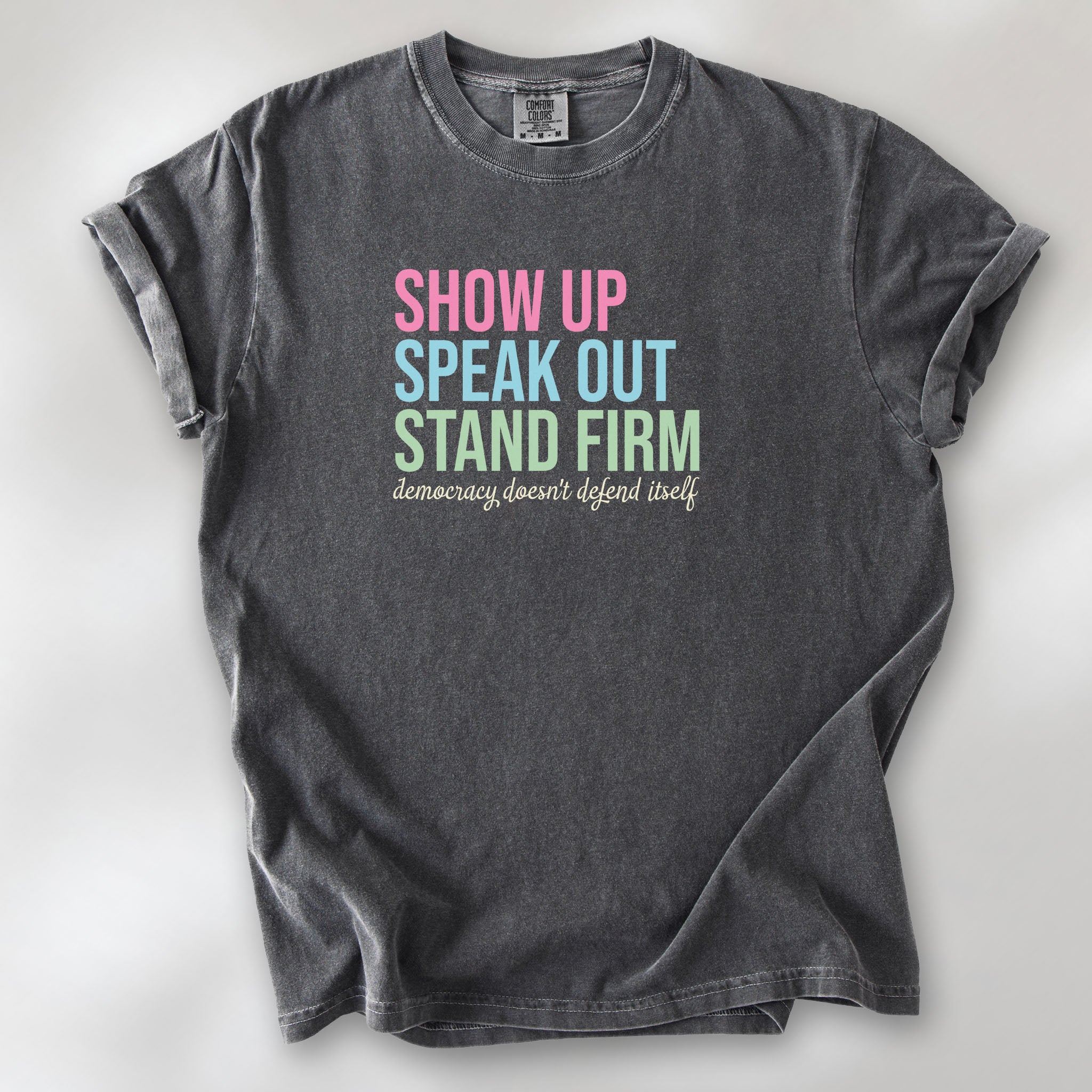 Pepper t-shirt with bright pink, blue and green bold text that says "show up, speak out, stand firm" and subtle grey script text below that reads "democracy doesn't defend itself."