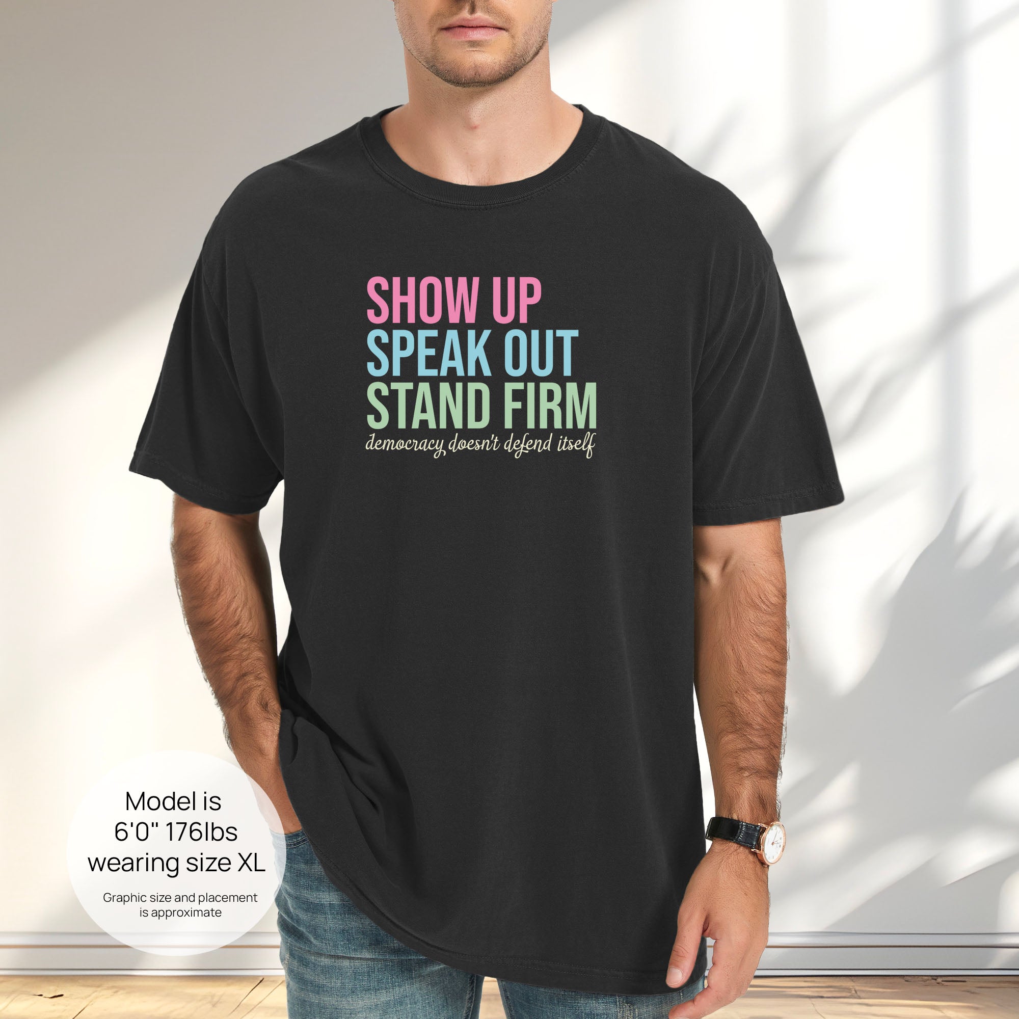 Male model wearing black t-shirt with bright pink, blue and green bold text that says "show up, speak out, stand firm" and subtle grey script text below that reads "democracy doesn't defend itself." Includes size and fit information for the model who is 6 feet 0 inches, 176 pounds, and wearing a size XL shirt.
