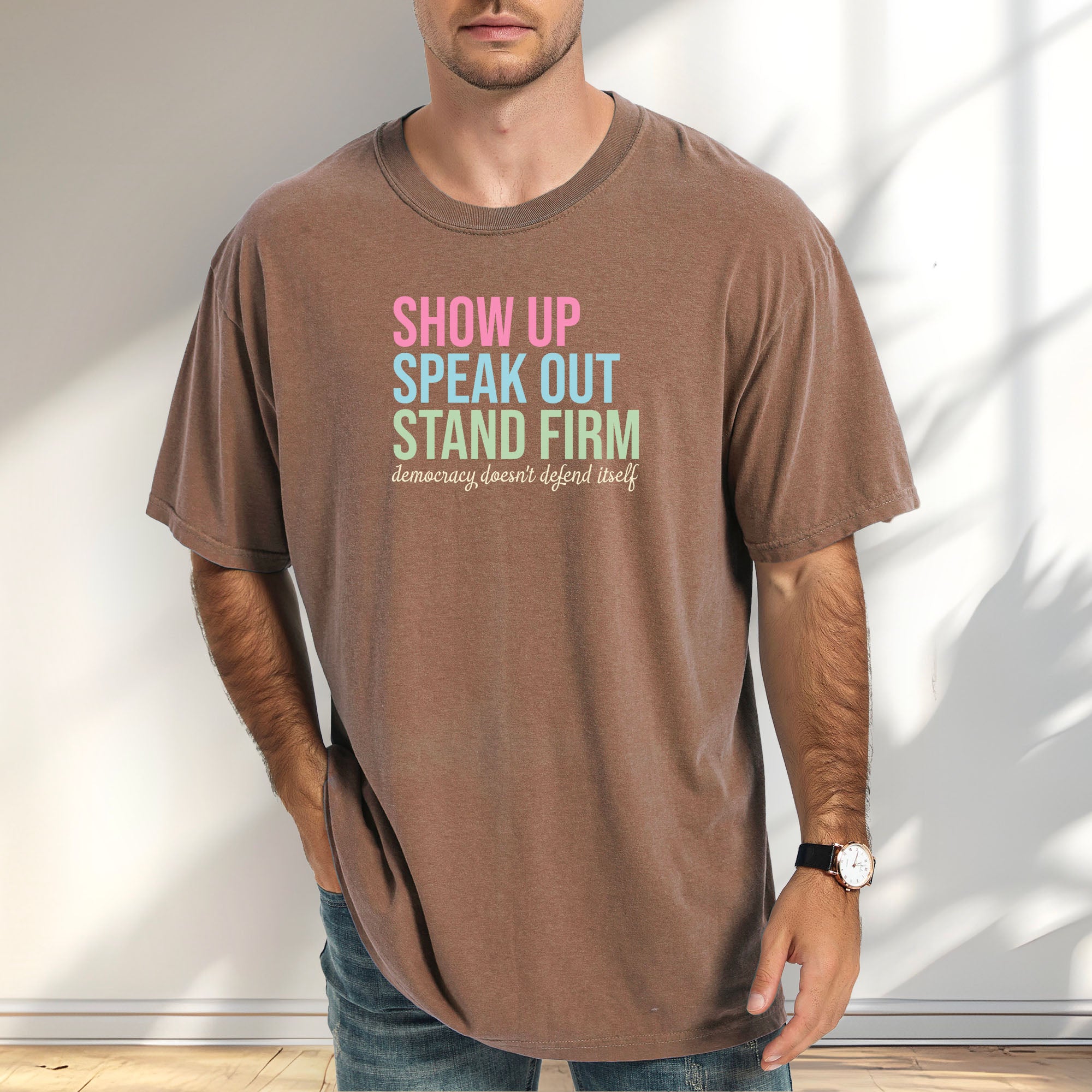Male model wearing espresso t-shirt with bright pink, blue and green bold text that says "show up, speak out, stand firm" and subtle grey script text below that reads "democracy doesn't defend itself."