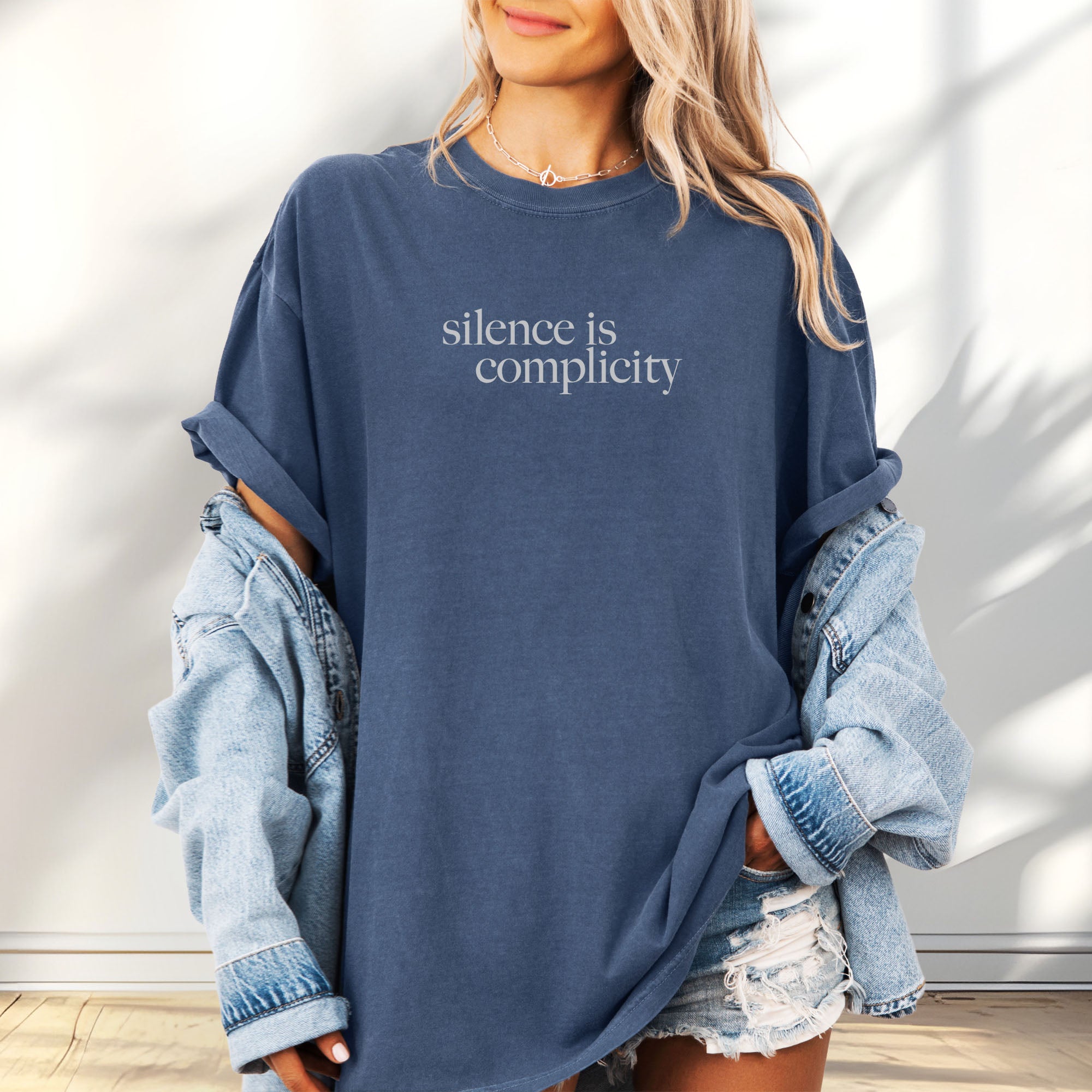 Female model wearing oversized denim t-shirt with minimalist serif text reading “silence is complicity."