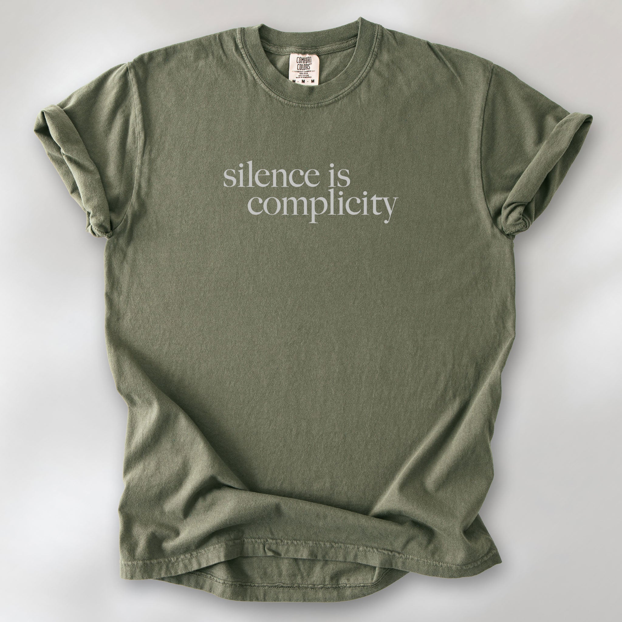 Sage t-shirt with minimalist serif text reading “silence is complicity."