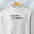 Hanging grey sweatshirt with minimalist serif text reading “silence is complicity."