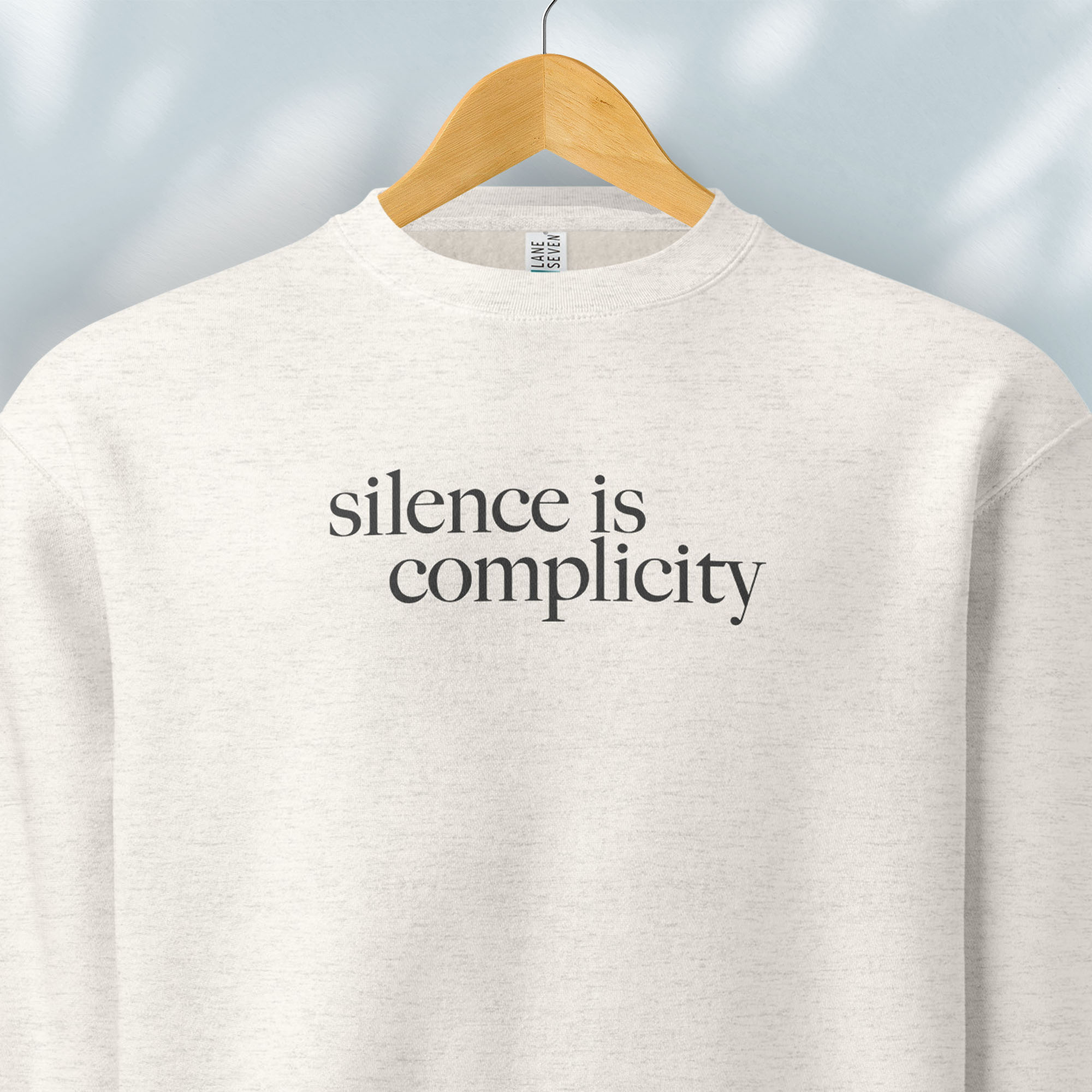 Hanging oatmeal sweatshirt with minimalist serif text reading “silence is complicity."