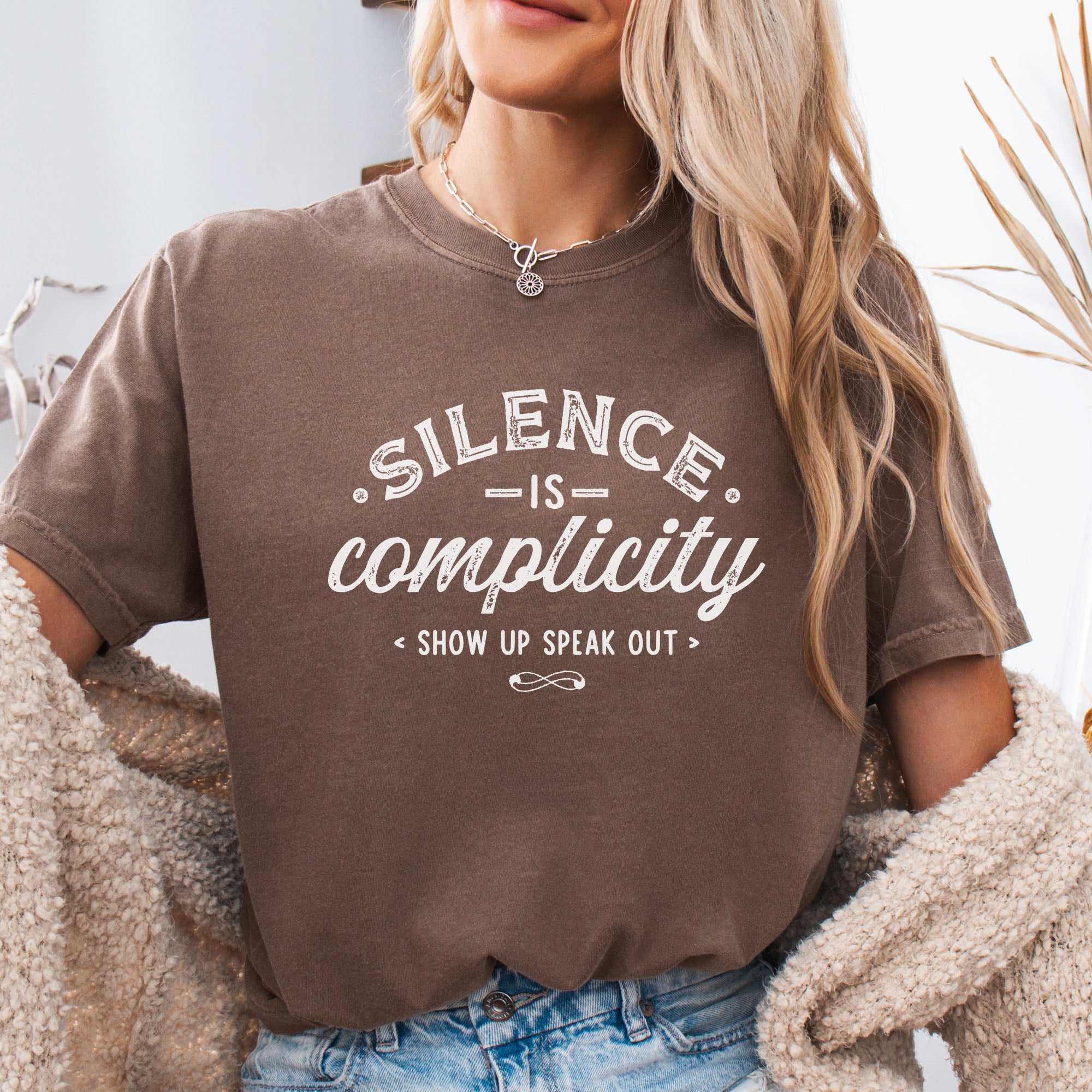 Female model wearing espresso t-shirt with vintage distressed typography reading “Silence Is Complicity – Show Up Speak Out.”