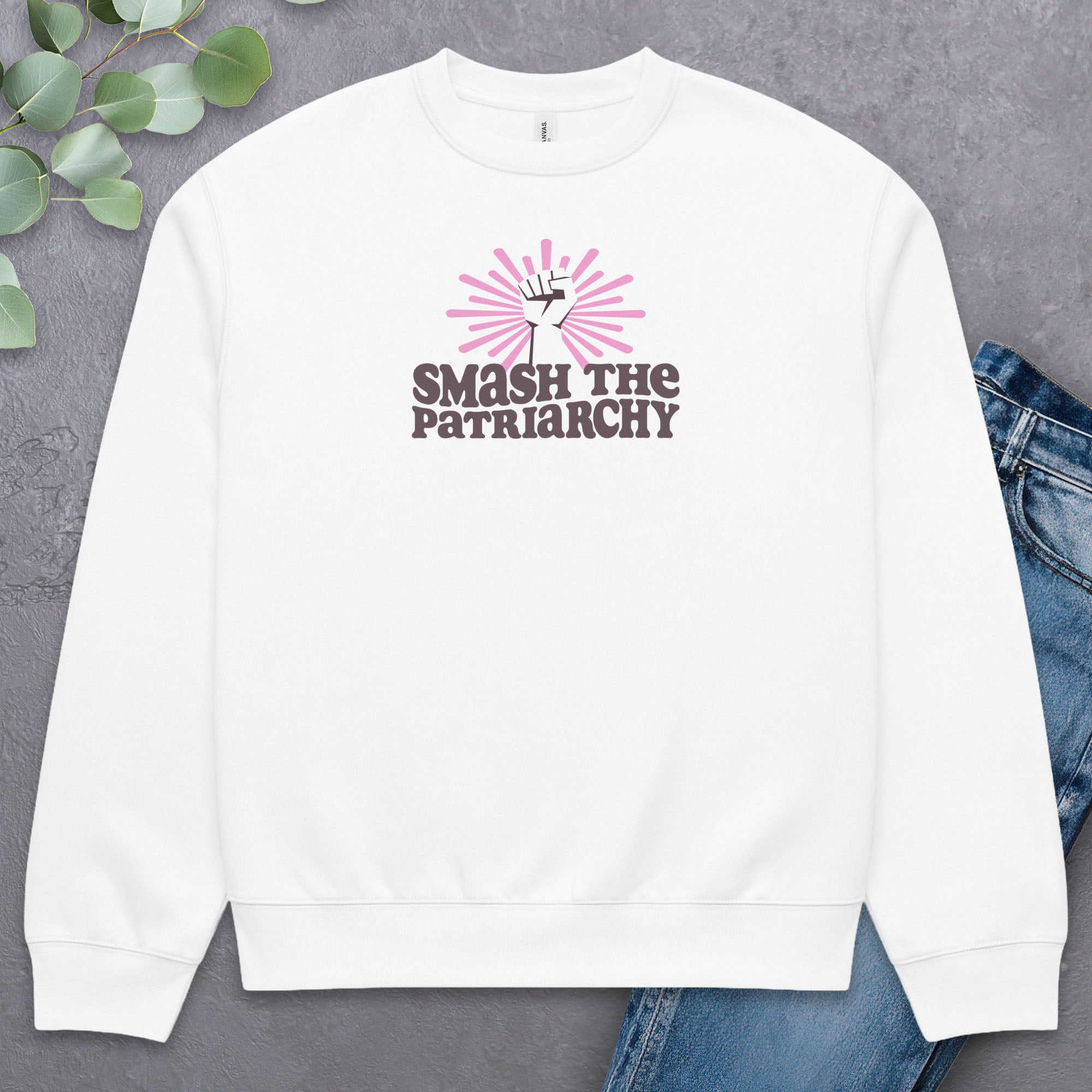 White sweatshirt with a raised fist graphic and pink rays behind it, featuring the text “Smash the Patriarchy.” Styled flat with a pair of jeans.