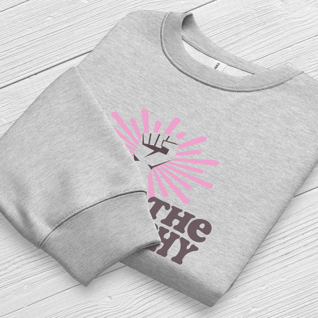 Folded athletic heather grey sweatshirt with a raised fist graphic and pink rays behind it, featuring the text “Smash the Patriarchy.” Shows cuff detail.