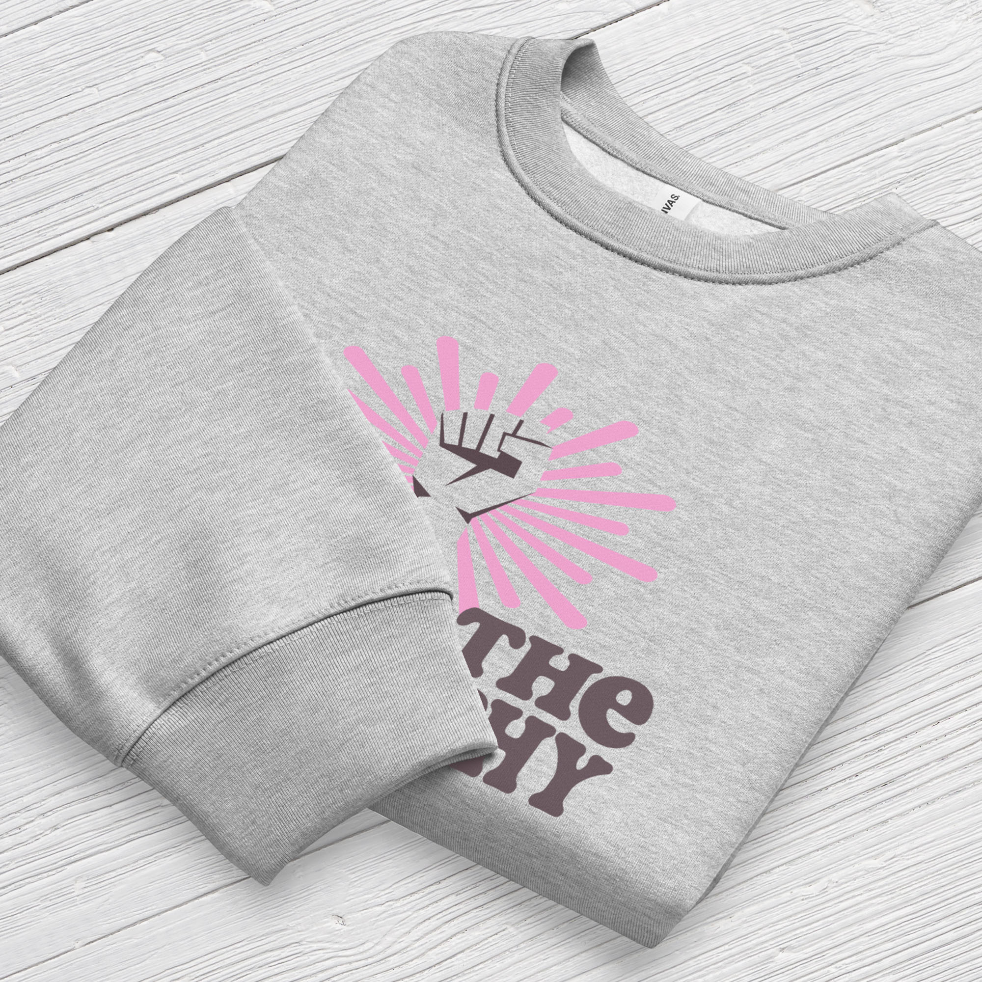 Folded athletic heather grey sweatshirt with a raised fist graphic and pink rays behind it, featuring the text “Smash the Patriarchy.” Shows cuff detail.