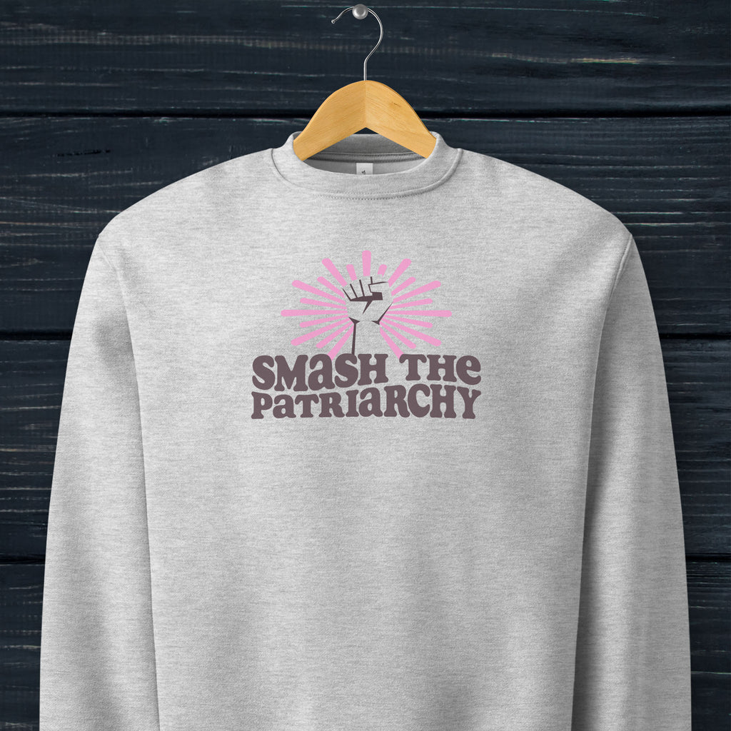 Grey sweatshirt with a raised fist graphic and pink rays behind it, featuring the text “Smash the Patriarchy.” Hanging on a hanger.