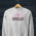 Grey sweatshirt with a raised fist graphic and pink rays behind it, featuring the text “Smash the Patriarchy.” Hanging on a hanger.