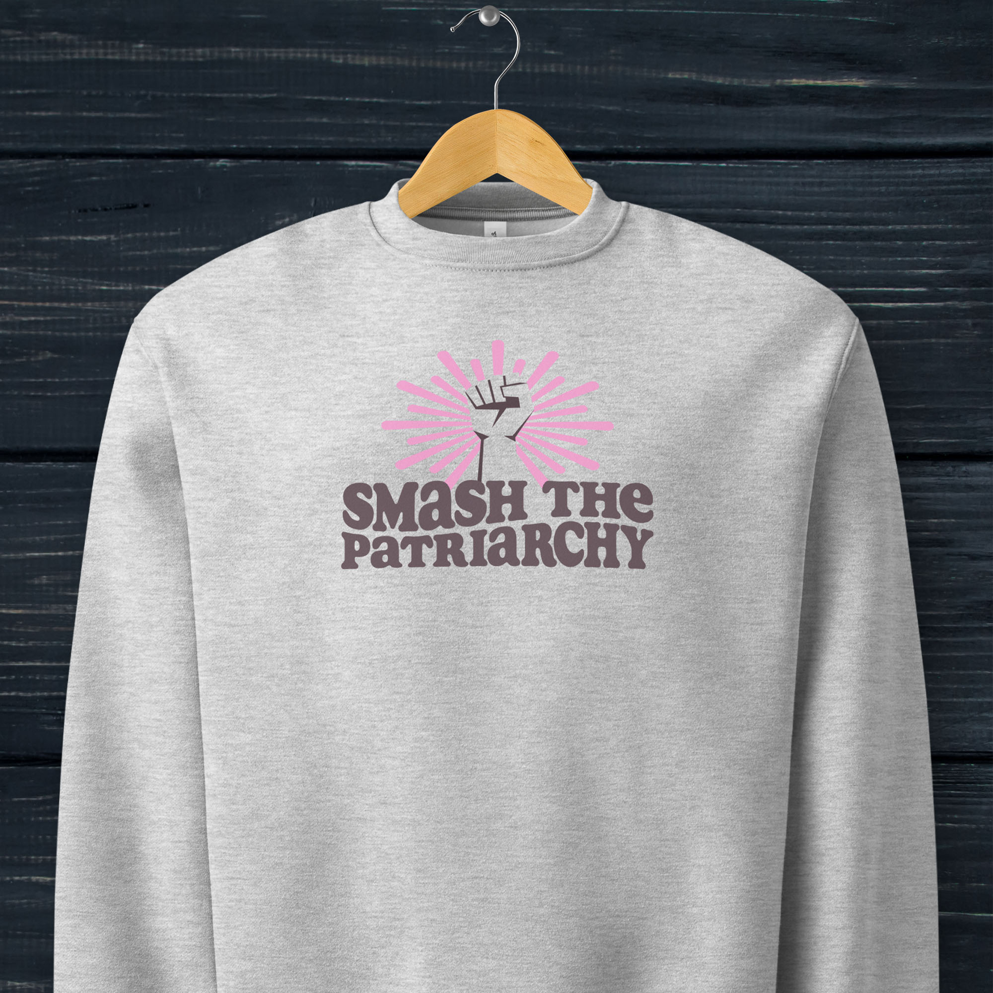 Grey sweatshirt with a raised fist graphic and pink rays behind it, featuring the text “Smash the Patriarchy.” Hanging on a hanger.